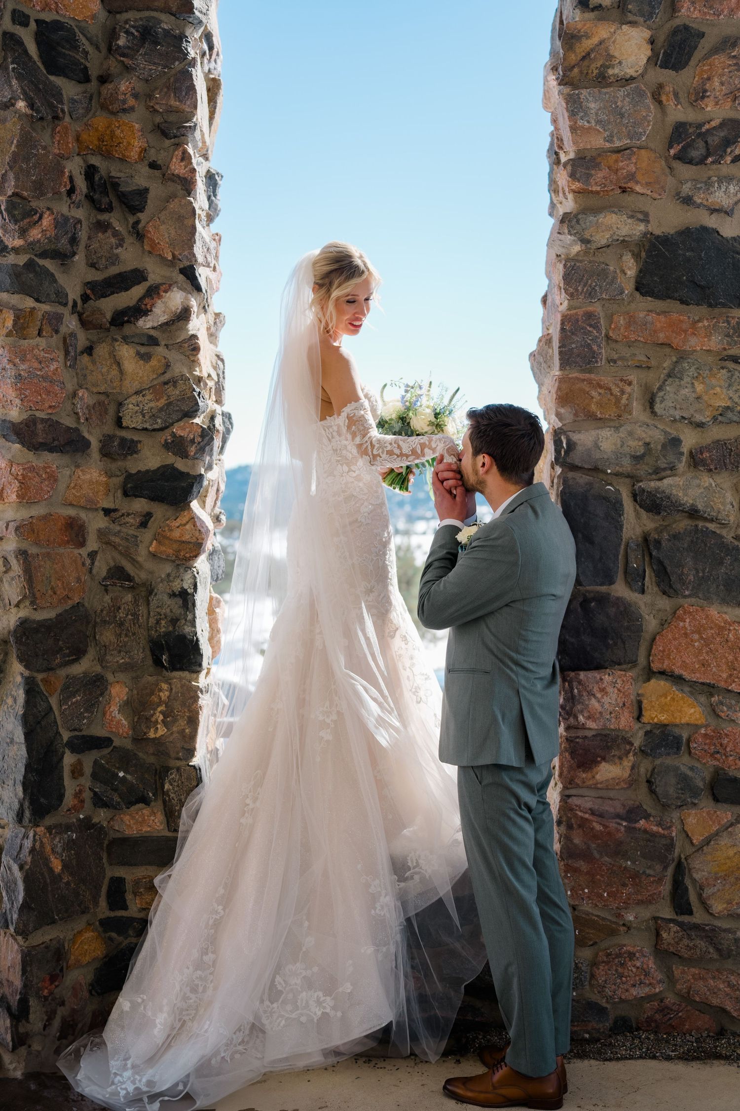 Bride in lace gown and veil holds bouquet as groom in gray suit kisses her hand by a rustic stone wall.