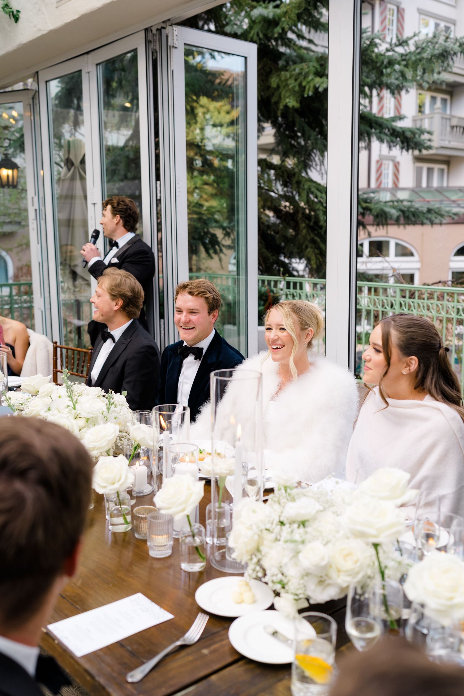 Bride and groom laughing at wedding reception head table adorned with white floral arrangements in elegant glass venue.