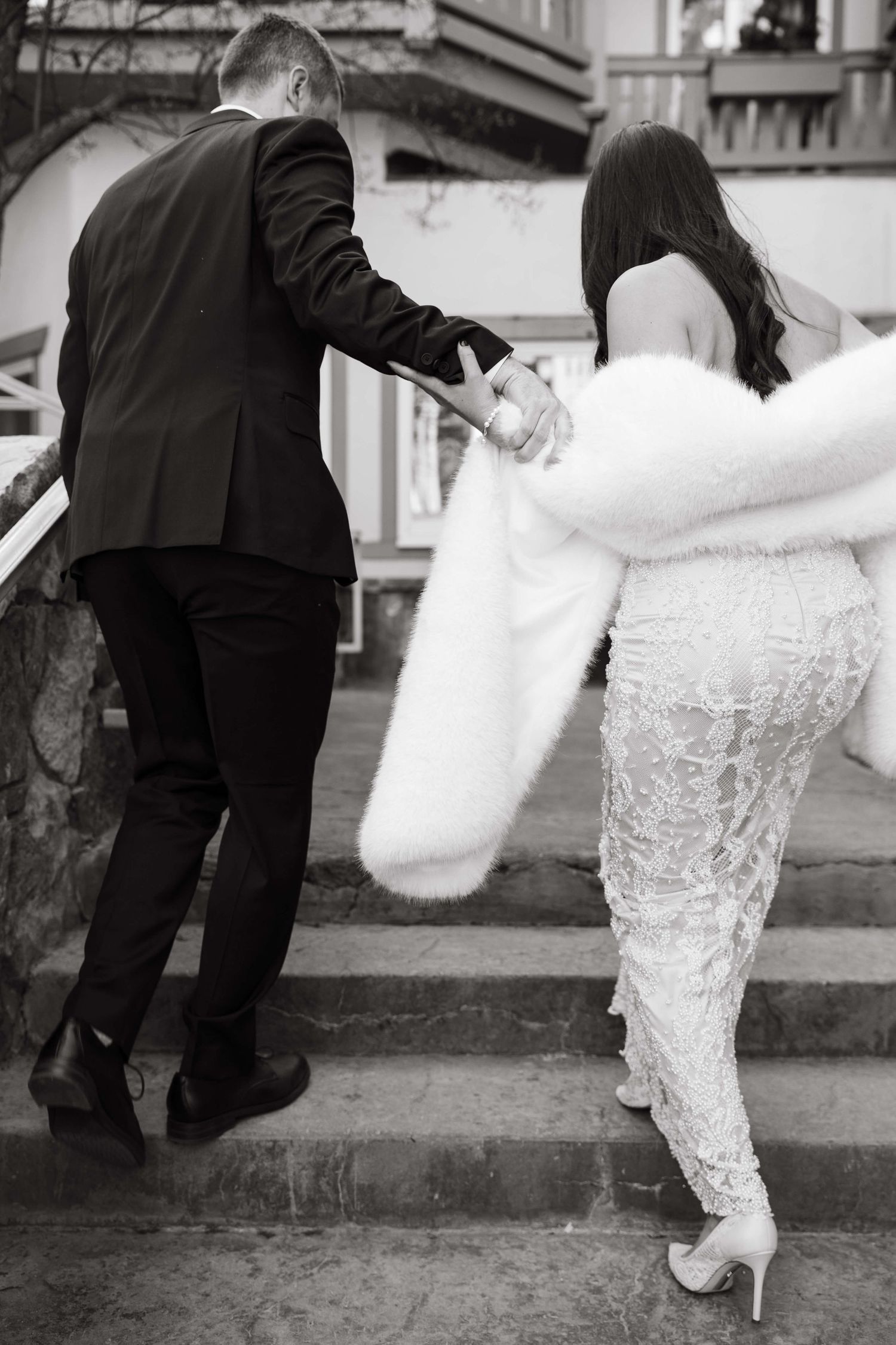 Black and white photo of bride and groom walking up stone steps, bride in lace gown with fur stole, holding hands.