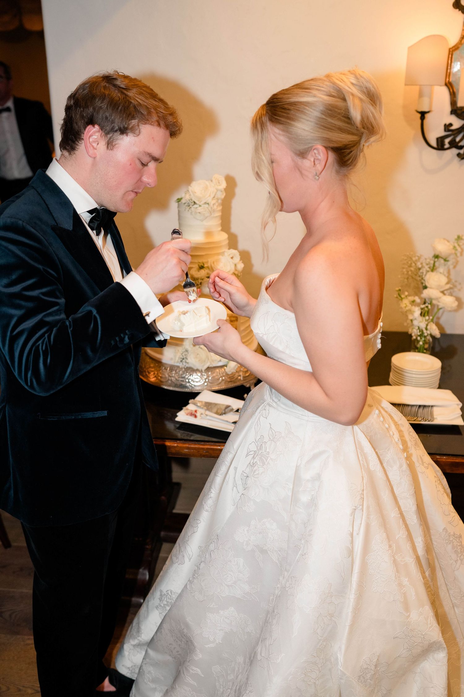 Bride and groom in black tuxedo cutting tiered wedding cake together at romantic indoor wedding reception.