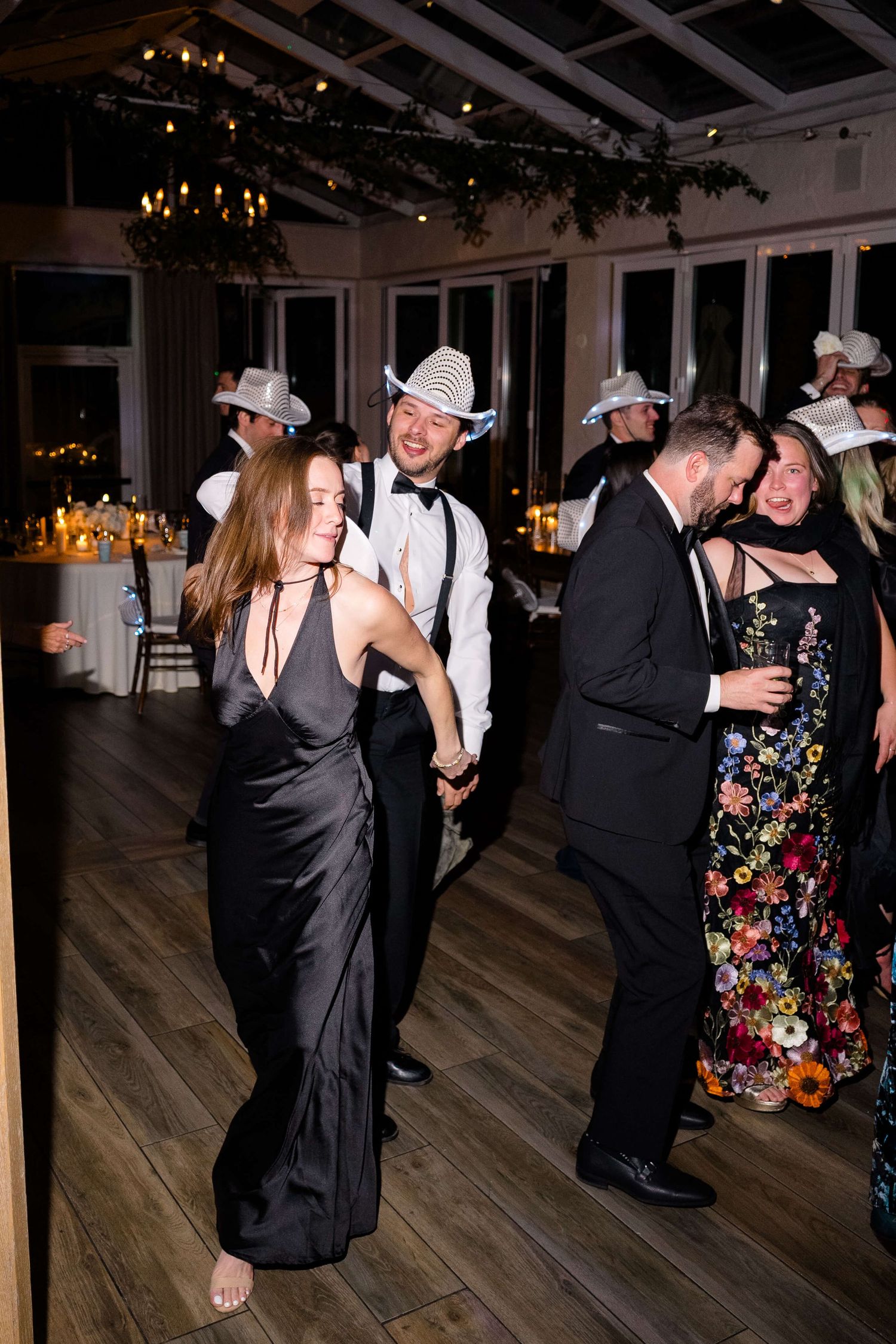 Guests dancing at an elegant wedding reception, wearing cowboy hats and formal attire in a candlelit venue.