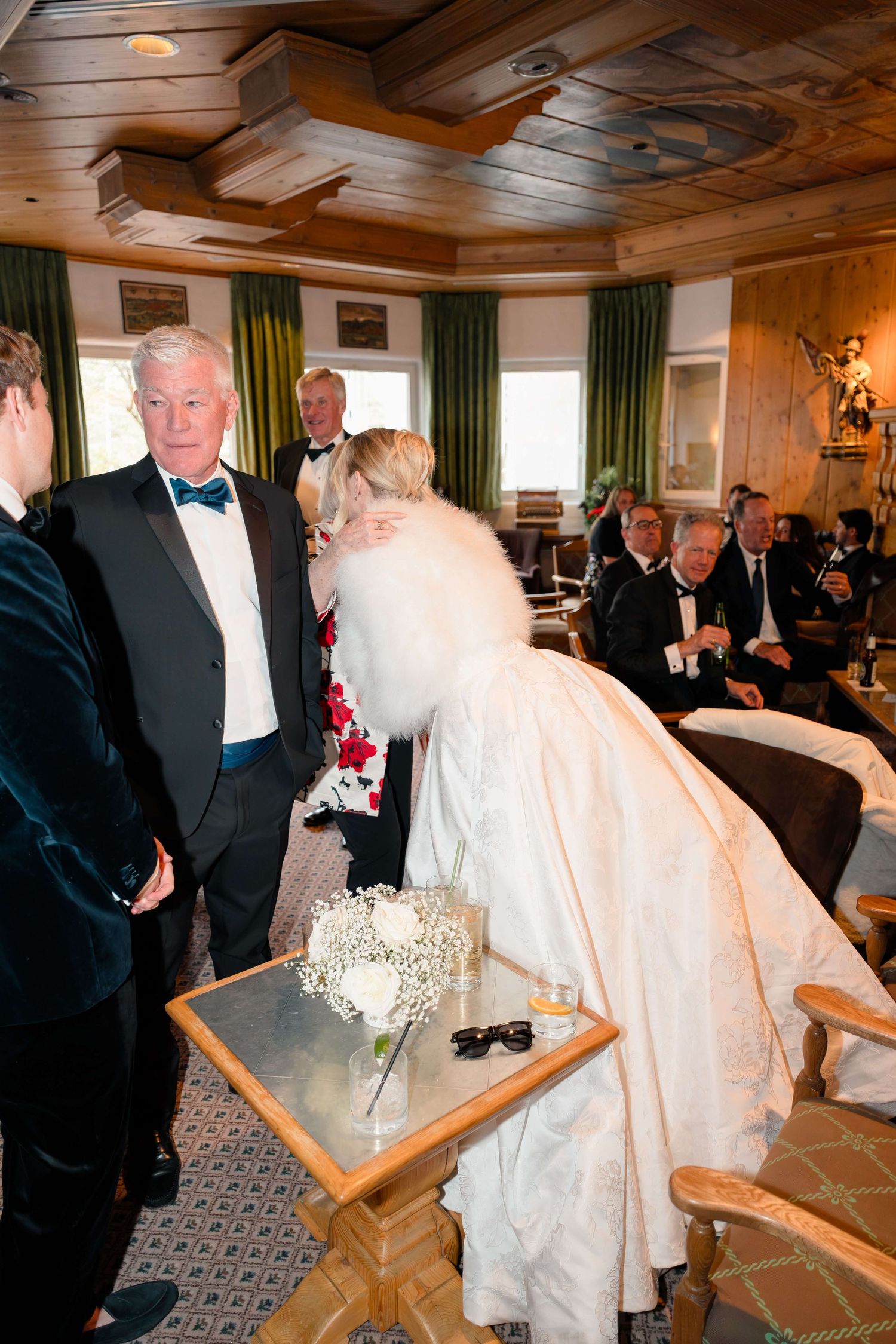 Bride in white gown leans over table with floral bouquet at elegant indoor wedding reception.