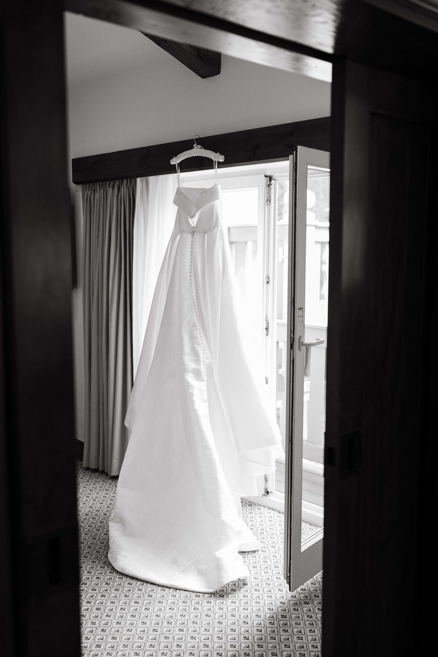 Black and white photo of an elegant bridal gown hanging in a hotel room doorway before a wedding ceremony.