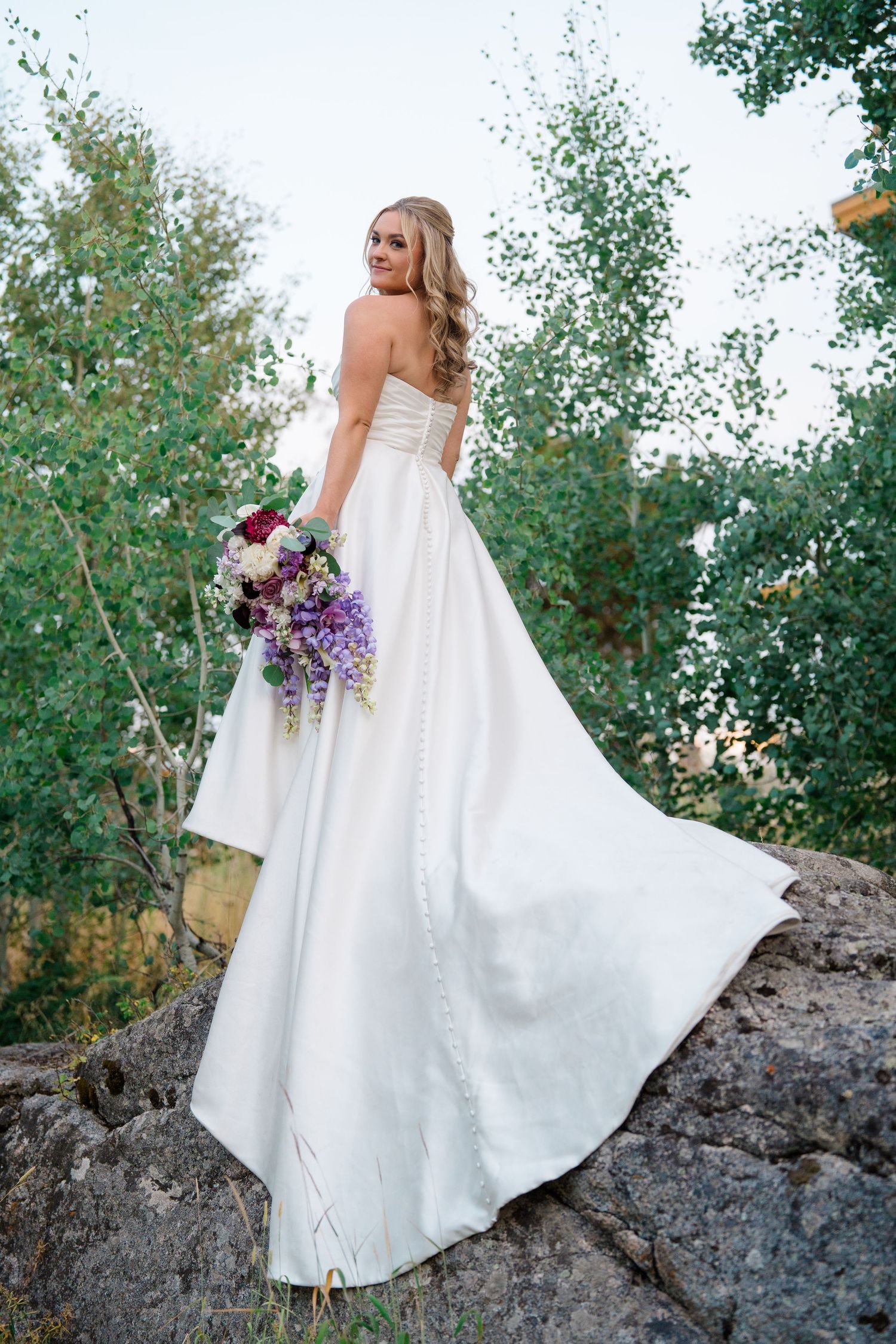 An elegant white wedding dress flowing in the breeze against a natural outdoor setting with rocks and trees.