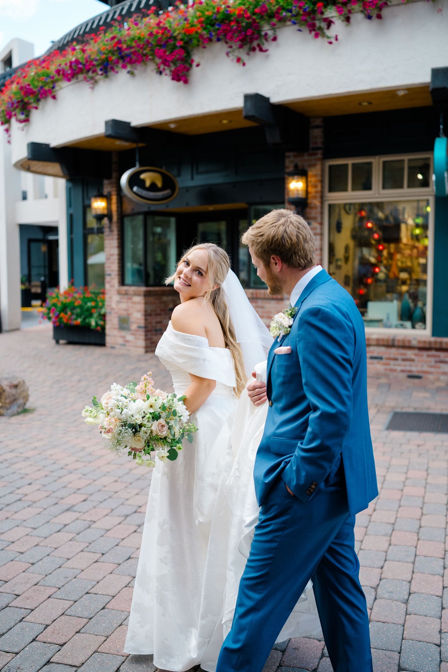 A couple in blue and white wedding attire walks along a brick pathway with hanging flowers overhead.