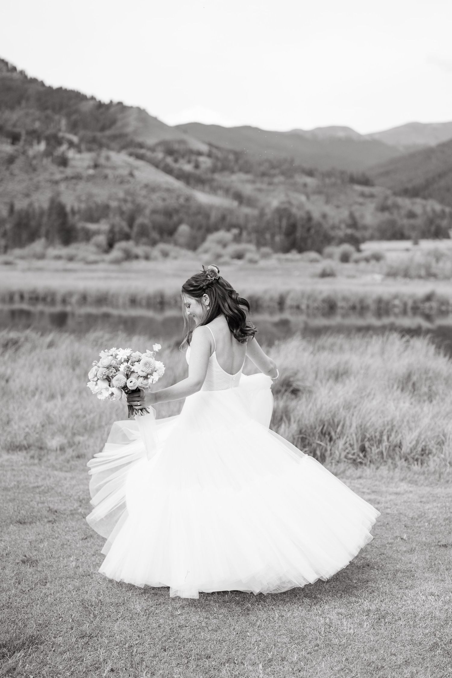 A bride in a flowing white dress twirls gracefully in a mountain meadow captured in black and white photography.