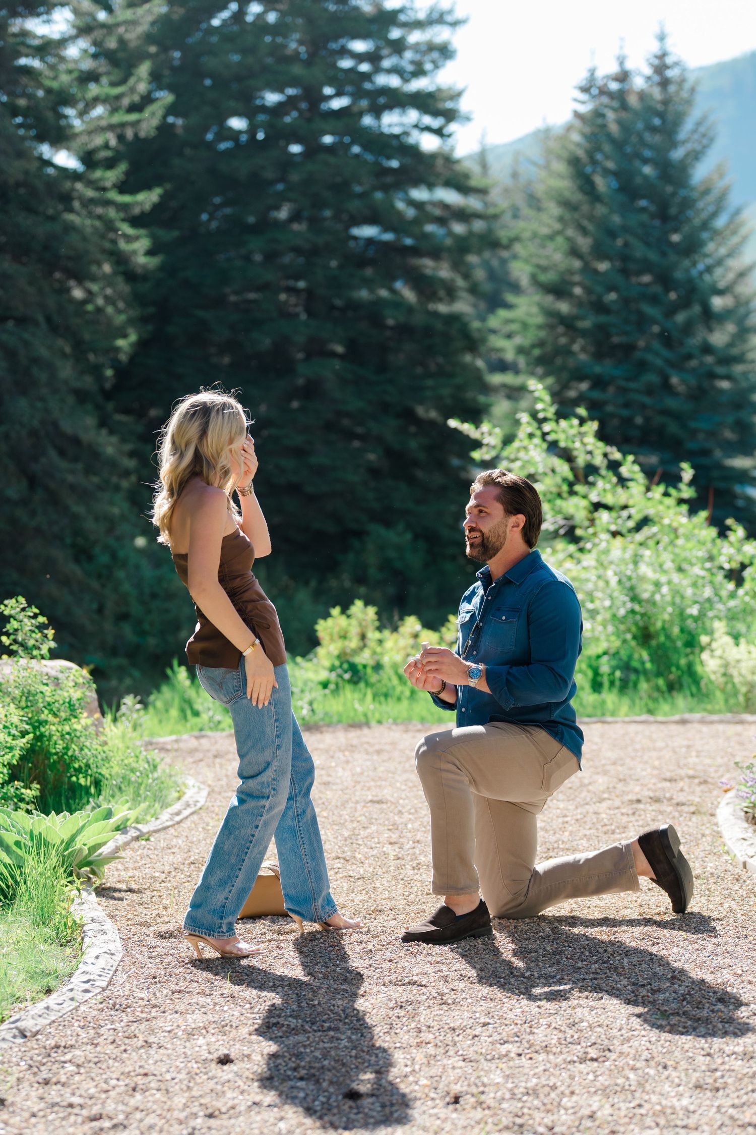 A couple shares a special moment during a surprise proposal on a scenic mountain trail with evergreen trees in the background.