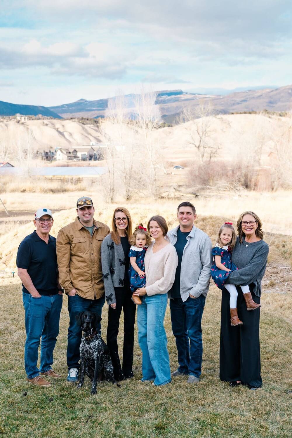 A group stands together outdoors on a grassy hill with mountains and bare winter trees in the background, vail family photograph locations