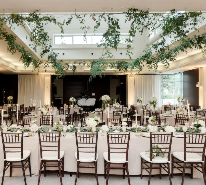 Elegant wedding reception hall with greenery ceiling installation, white linens, chiavari chairs, and floral centerpieces.