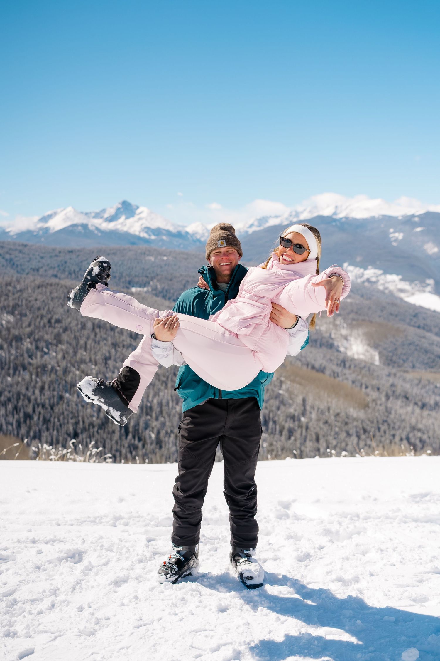 Two people playfully pose in winter snow gear against a stunning mountain backdrop with snow-covered peaks.