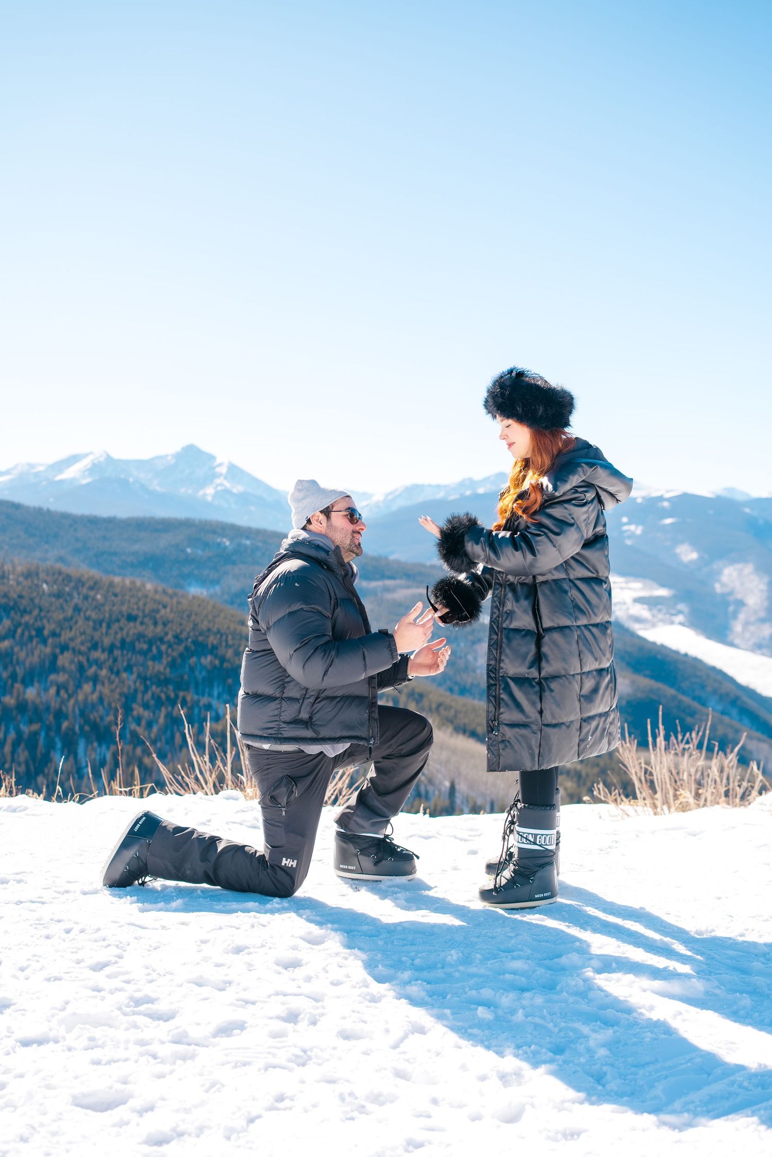 Two people sit together in winter coats enjoying hot drinks on a snowy mountain overlook with scenic mountain views.