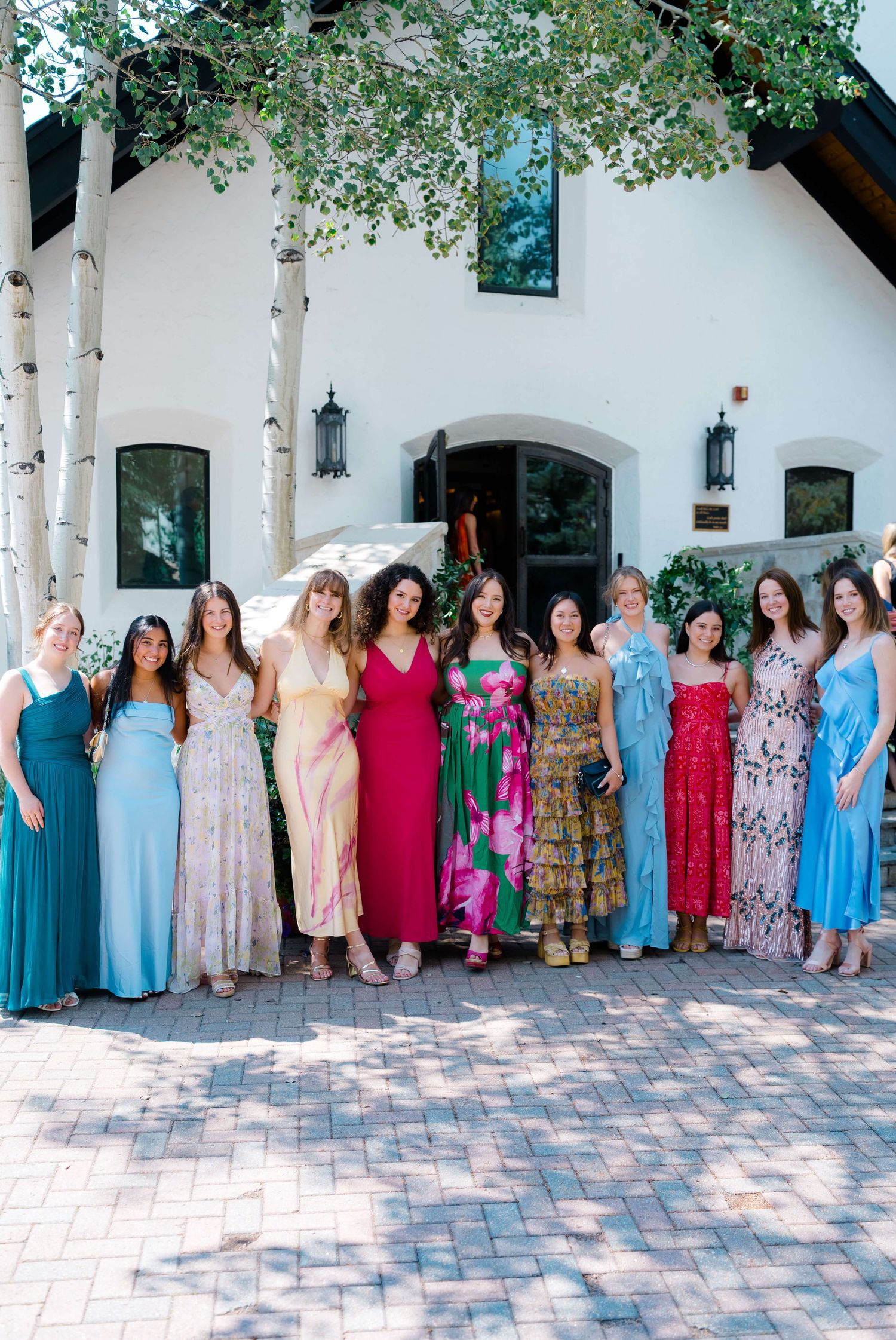 A panoramic photo of a group wearing colorful summer dresses lined up against a white building with arches and trees during vail wedding.