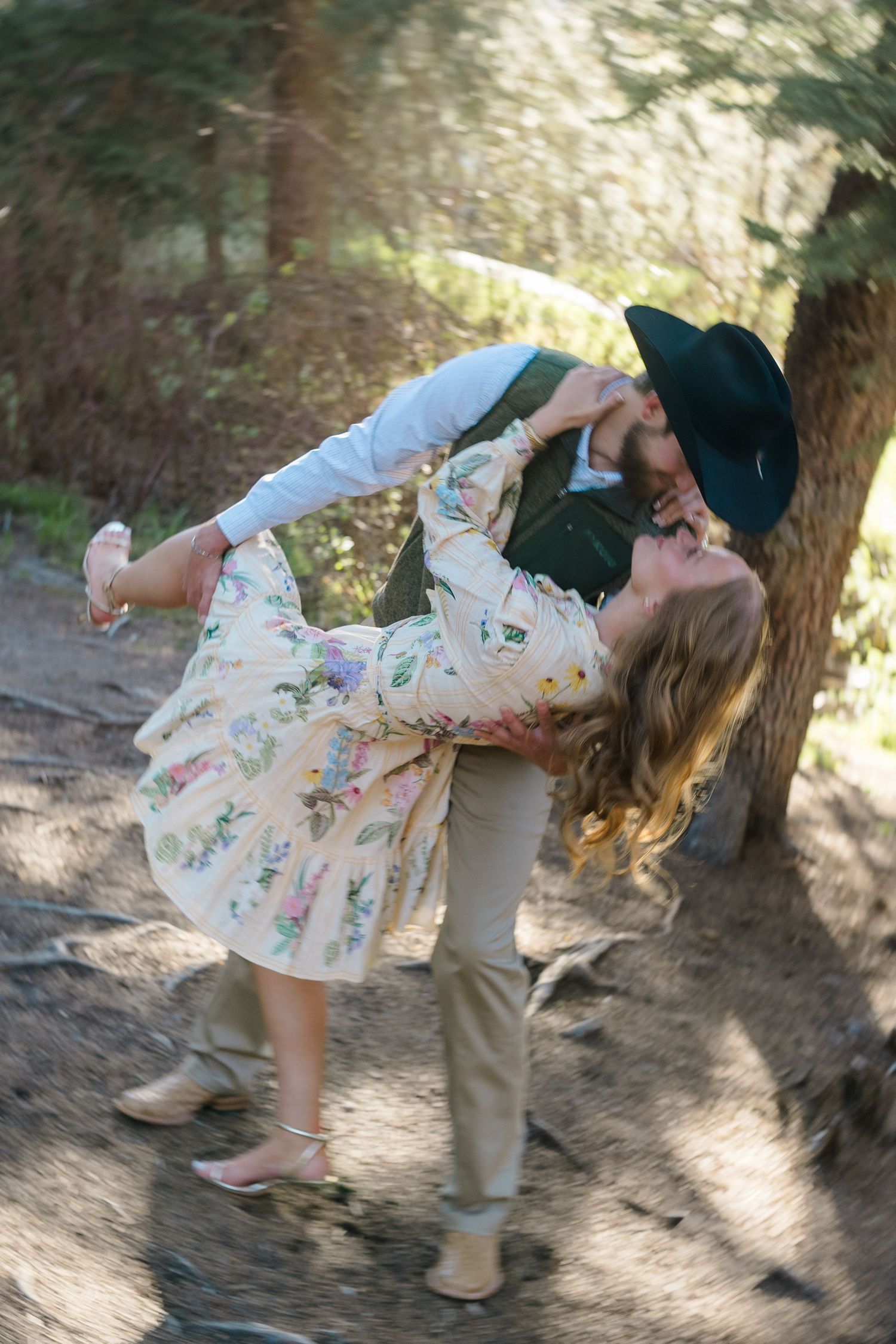 Couple sharing a romantic kiss outdoors, man in green hat lifting woman in floral dress near a sunlit tree.
