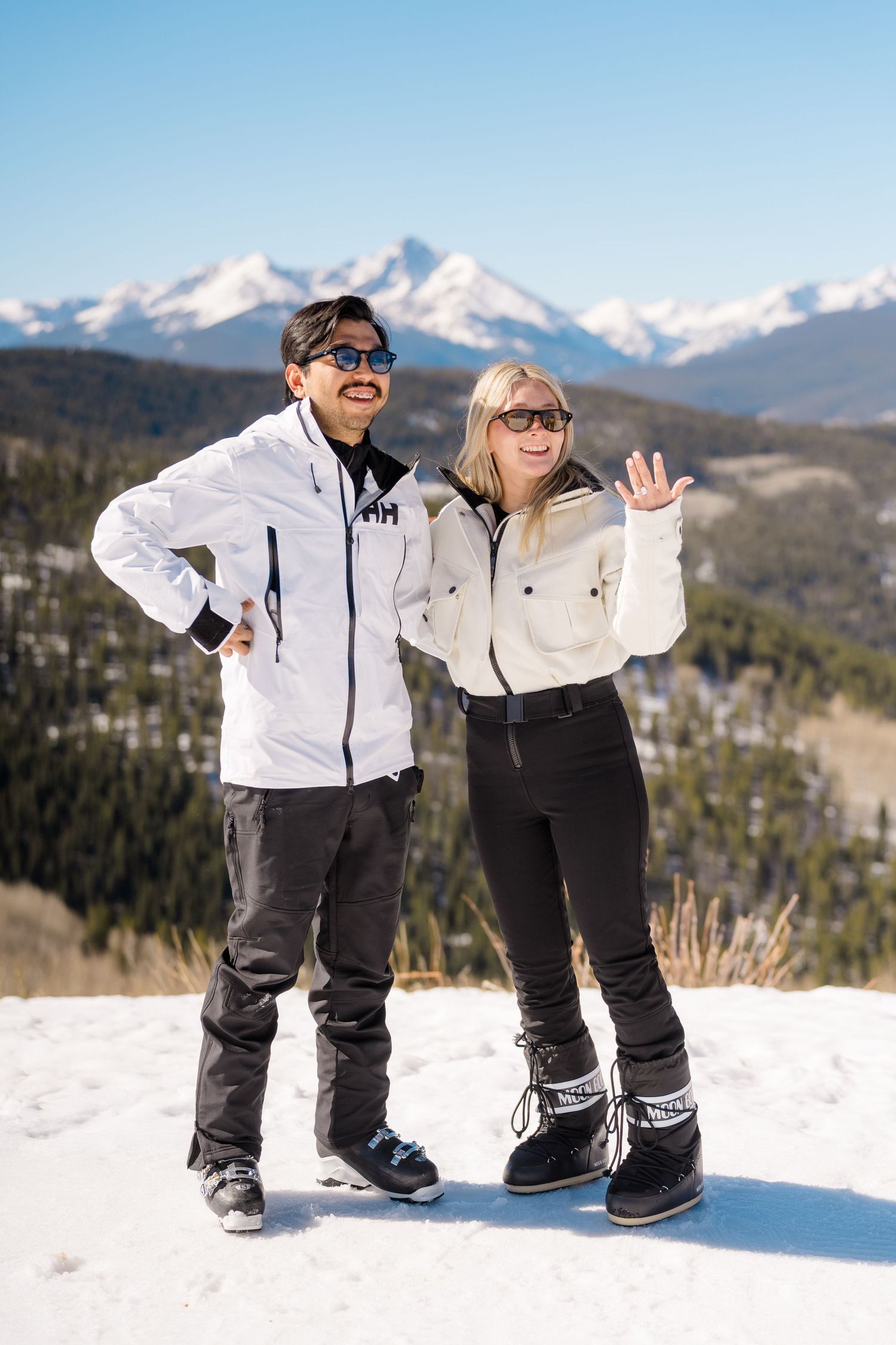 Two people in ski gear pose on a snowy mountain summit with scenic snow-capped peaks and blue sky behind them.