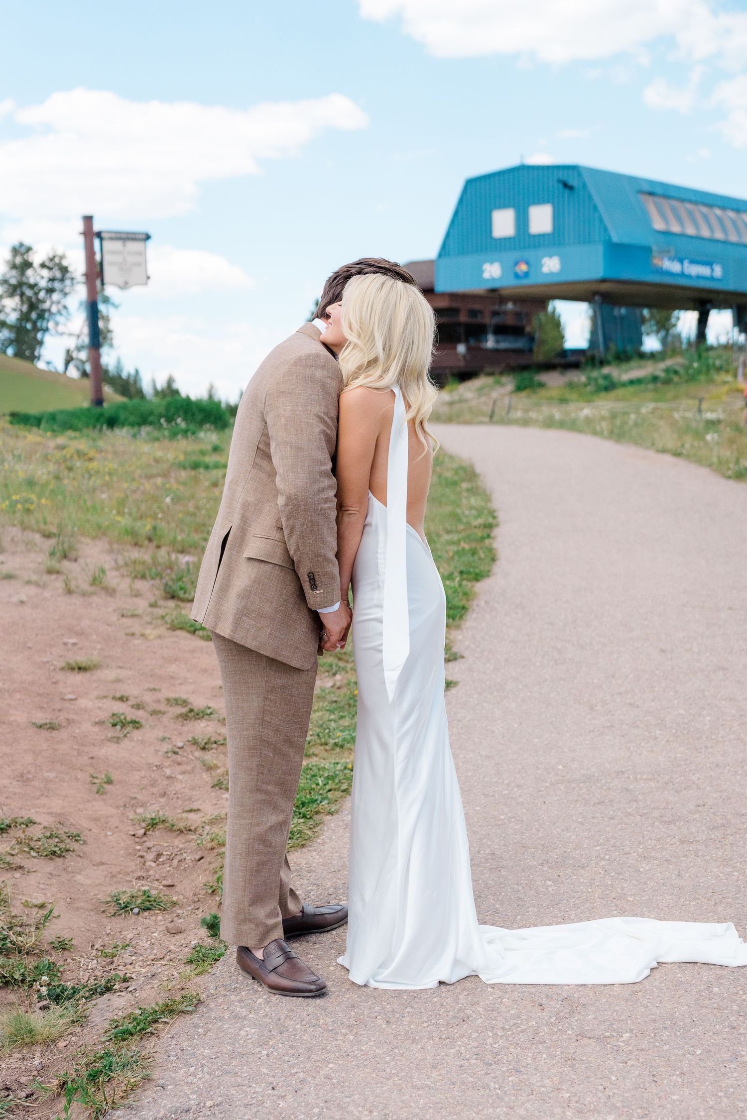 Bride and groom kissing on a mountain path, bride in white backless gown, groom in tan suit, ski lodge in background.