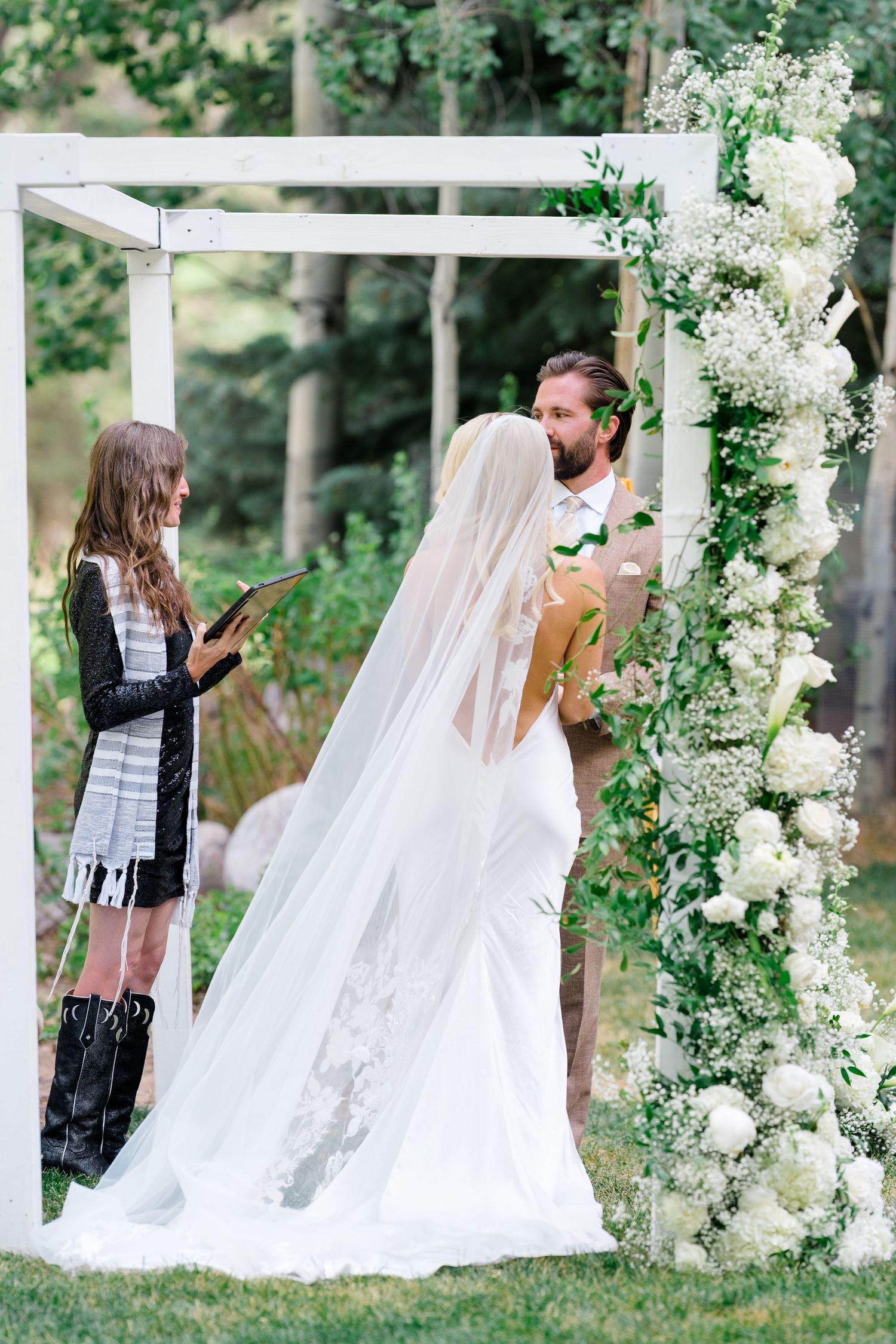 Bride and groom exchange vows under a white floral arch adorned with greenery and white roses at an outdoor wedding.