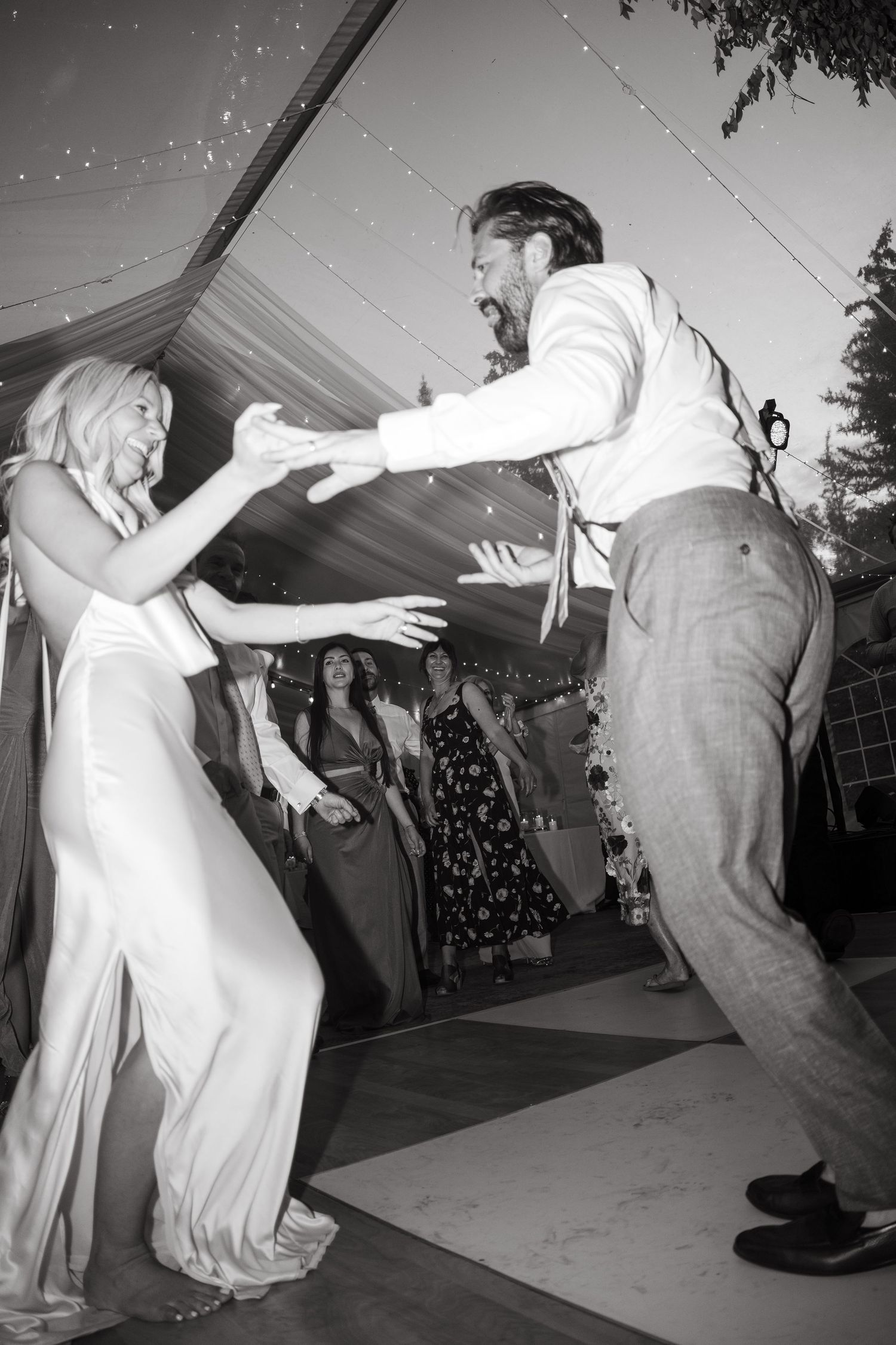 Black and white photo of bride and groom dancing energetically at outdoor wedding reception under tent lights.