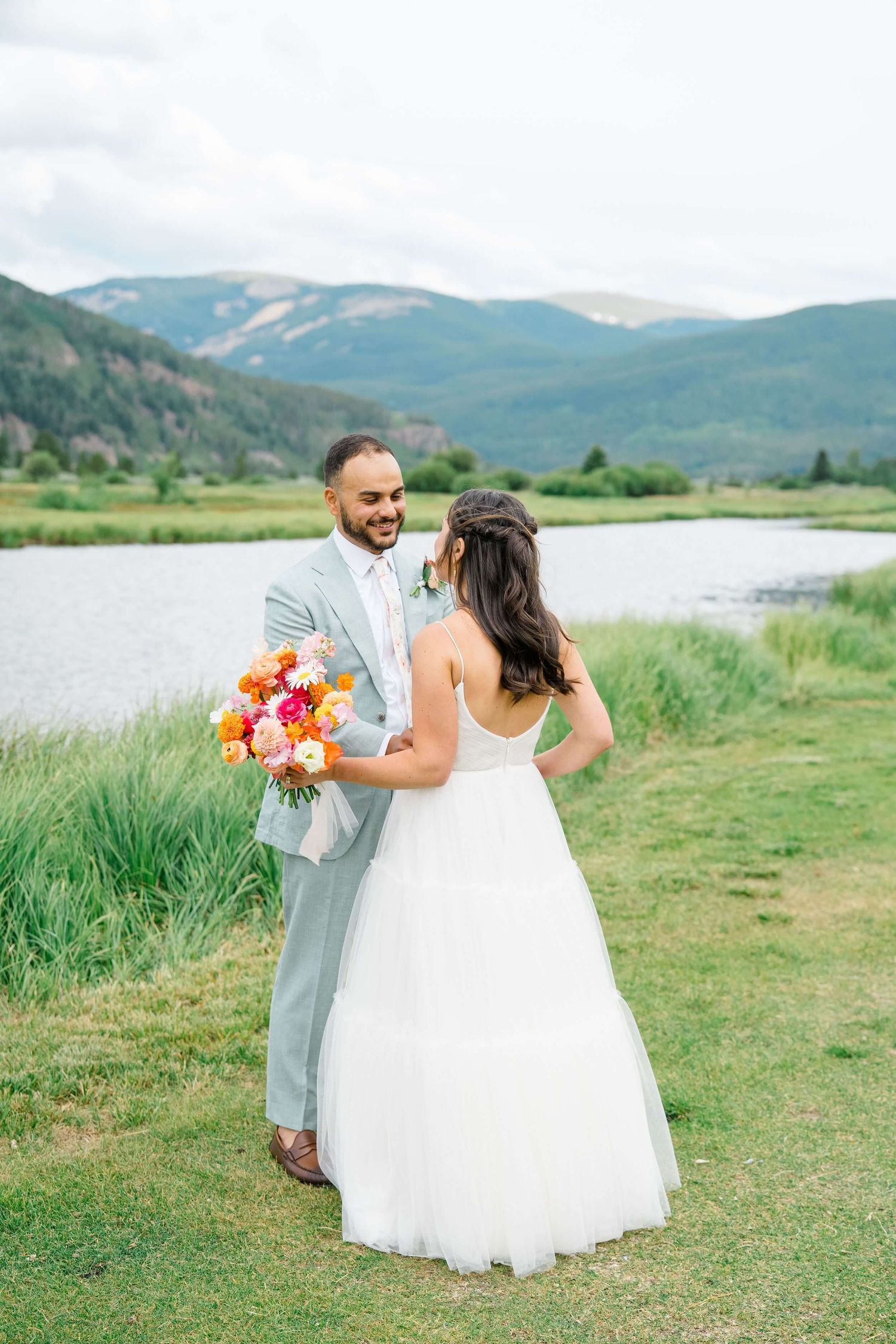 A couple in wedding attire dances joyfully on grassy meadow with mountain lake and evergreen forest backdrop at Camp Hale.