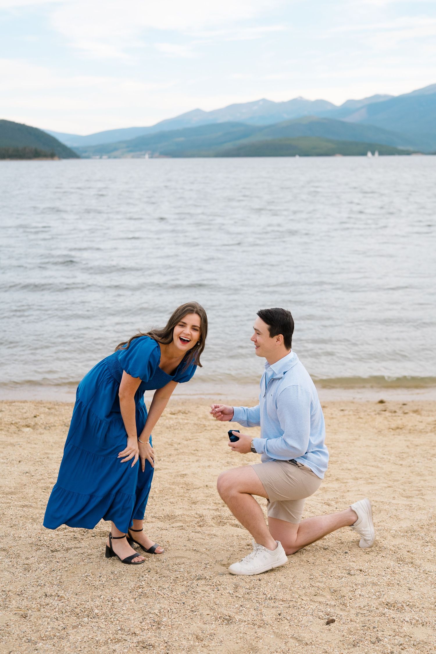A couple shares a romantic proposal moment on a sandy beach with mountains and lake in the background.