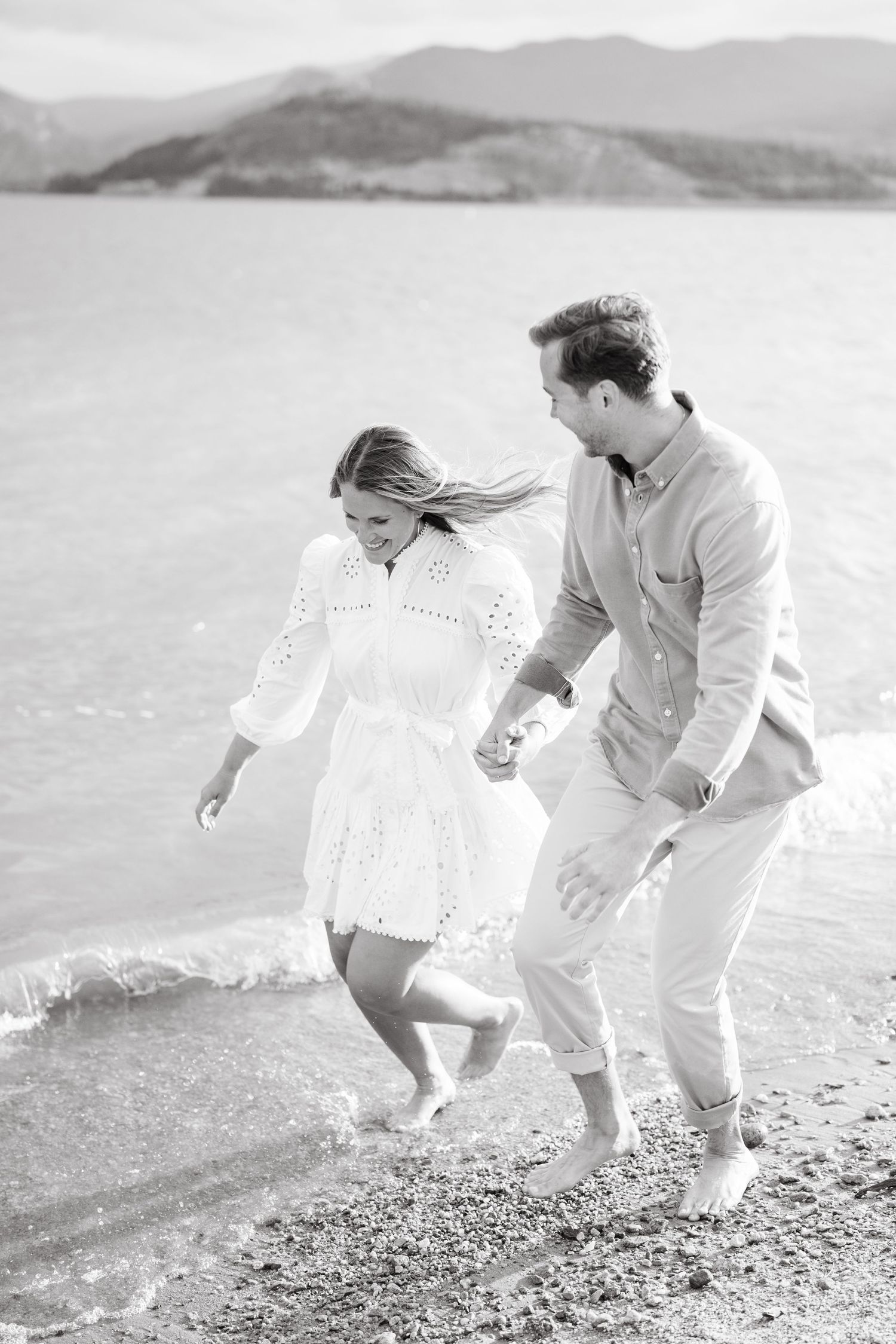 A couple in white outfits playfully runs along a scenic beach shoreline in black and white photography.