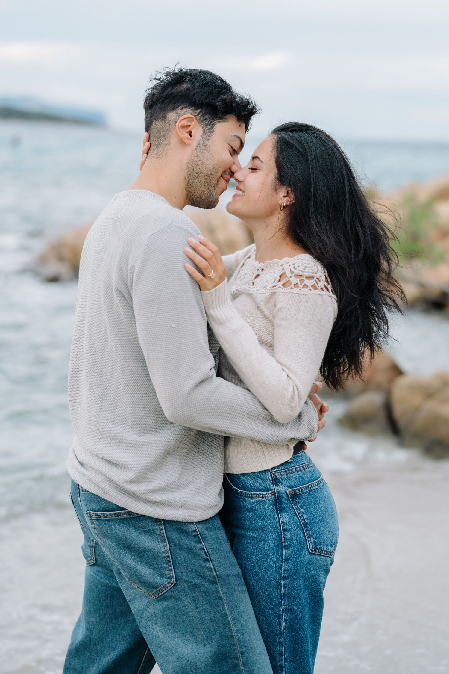 A romantic couple embraces and kisses on a rocky beach shoreline wearing jeans and casual attire.