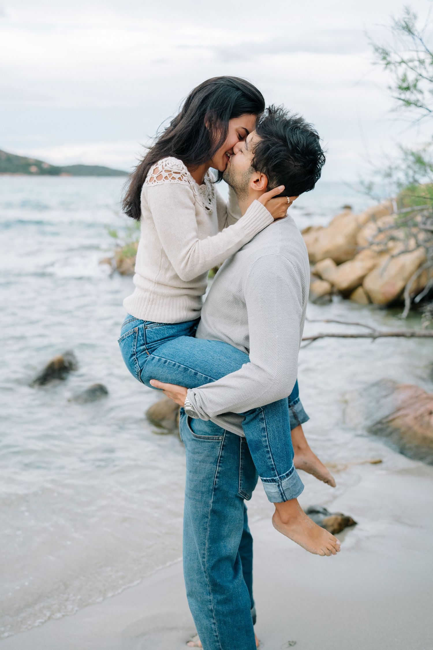 Romantic couple embracing on rocky beach shore with ocean waves and cloudy sky in background.
