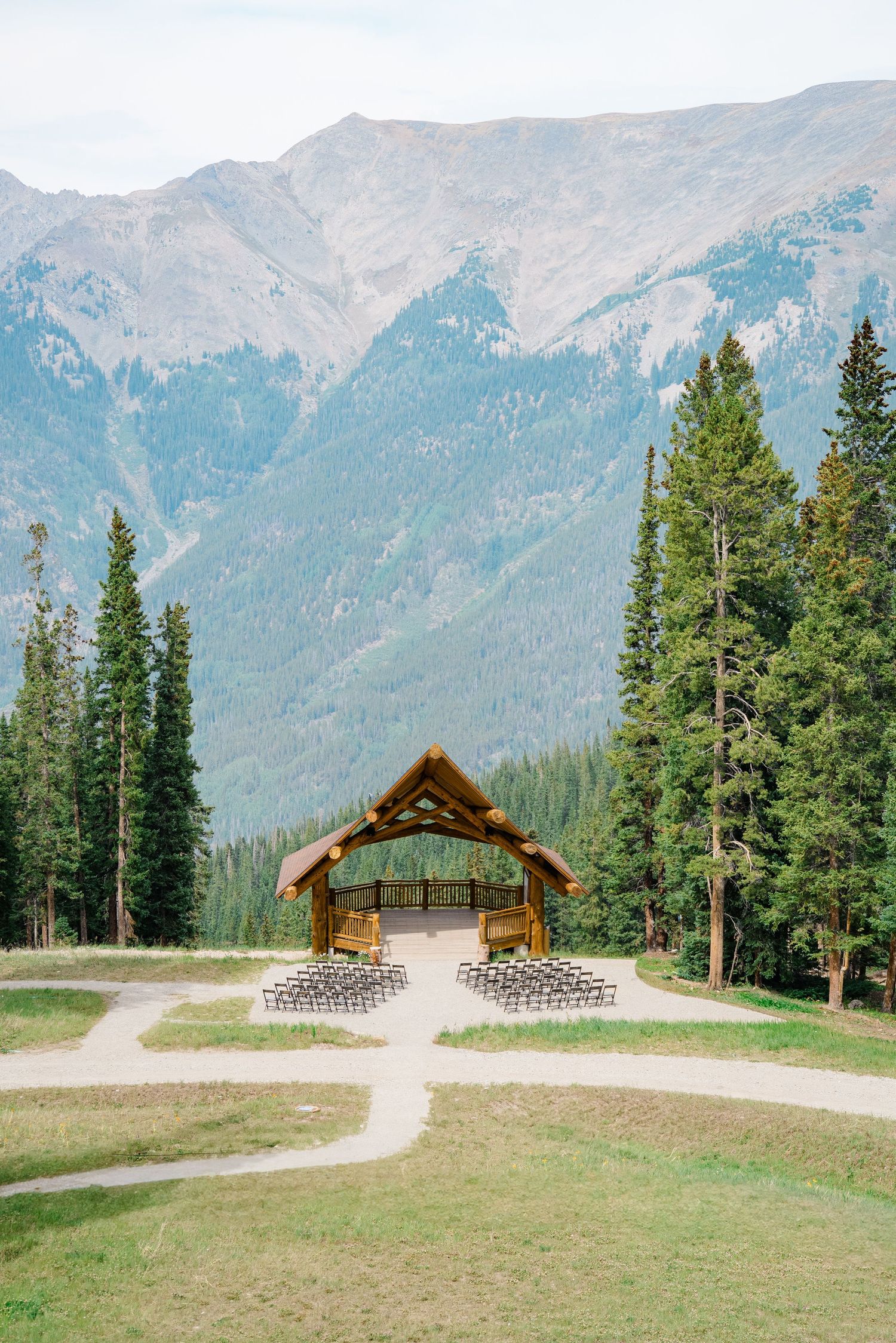 A rustic wooden wedding arch stands in a mountain meadow surrounded by evergreen trees and towering peaks in the background.