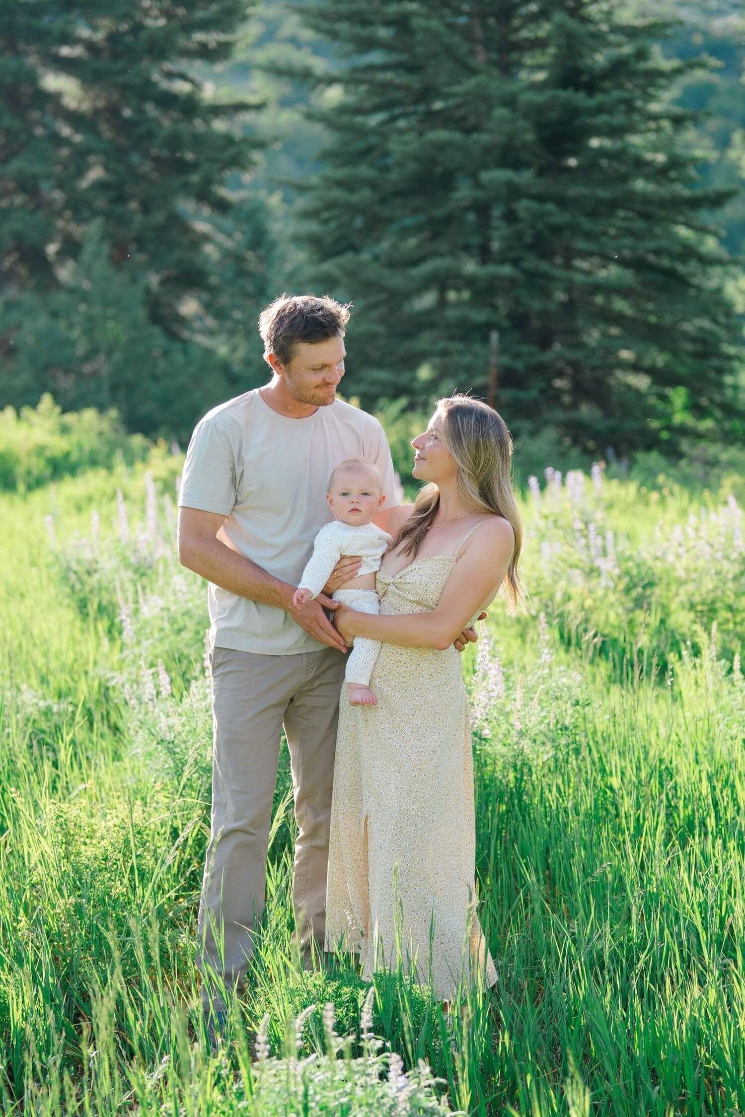 A family stands together in a meadow of wildflowers with evergreen trees in the background on a sunny day.