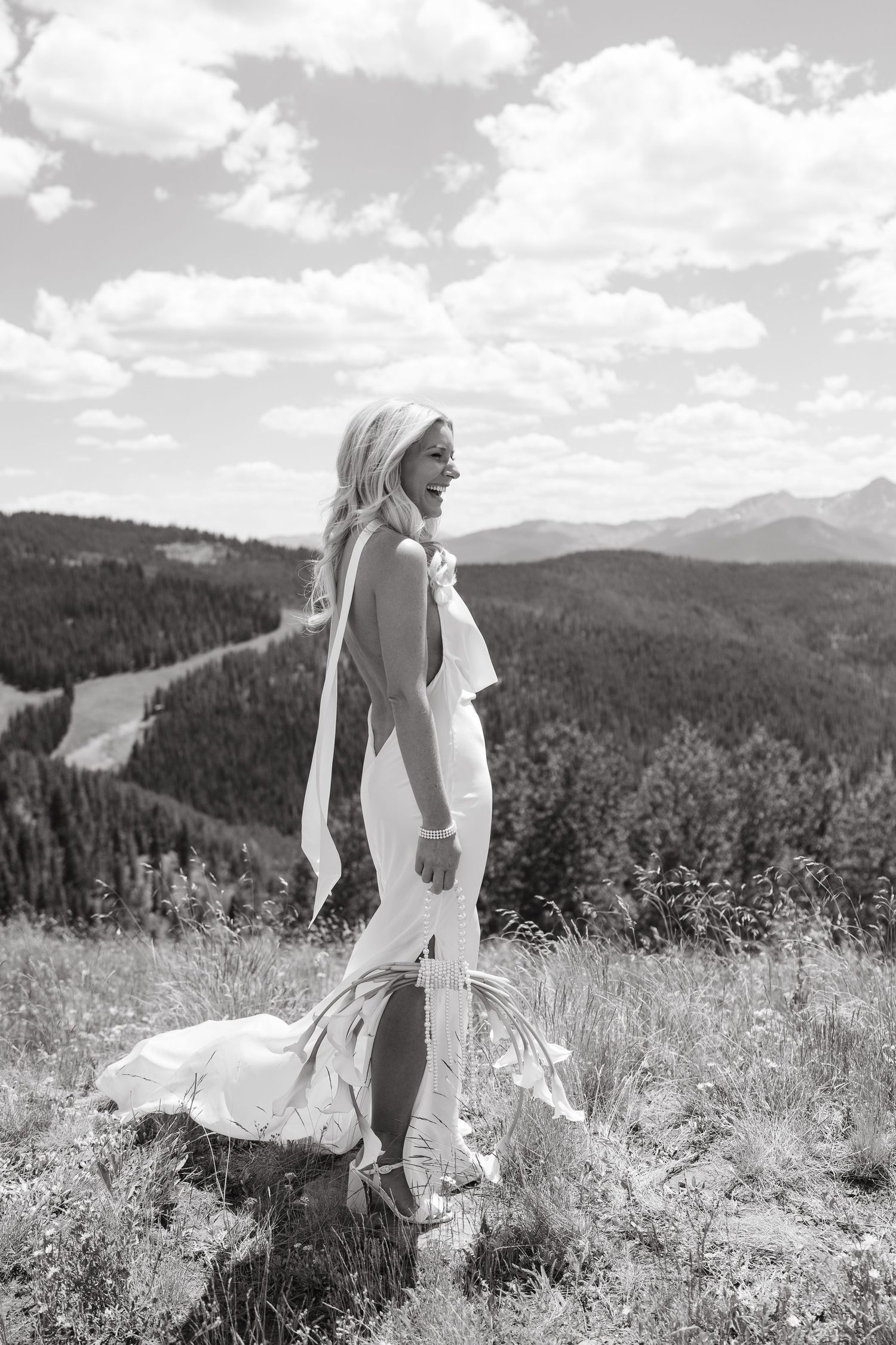 Black and white photo of a smiling bride in a white gown standing on a mountain meadow with forest and peaks behind her.