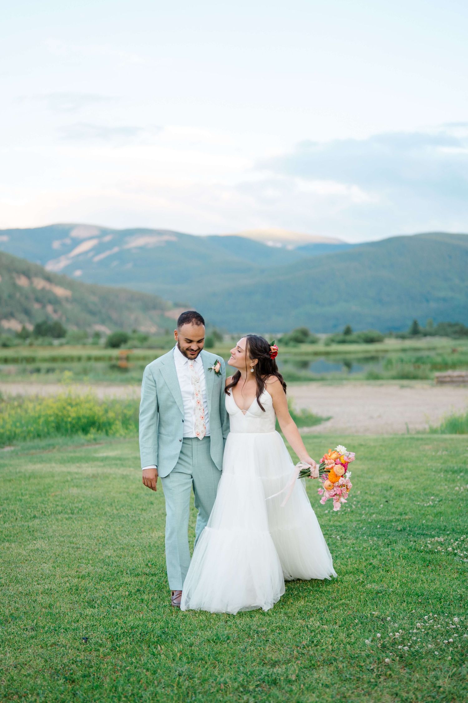 At Camp Hale Wedding Venue - Bride and groom walking hand in hand on green grass with stunning mountain backdrop at golden hour.