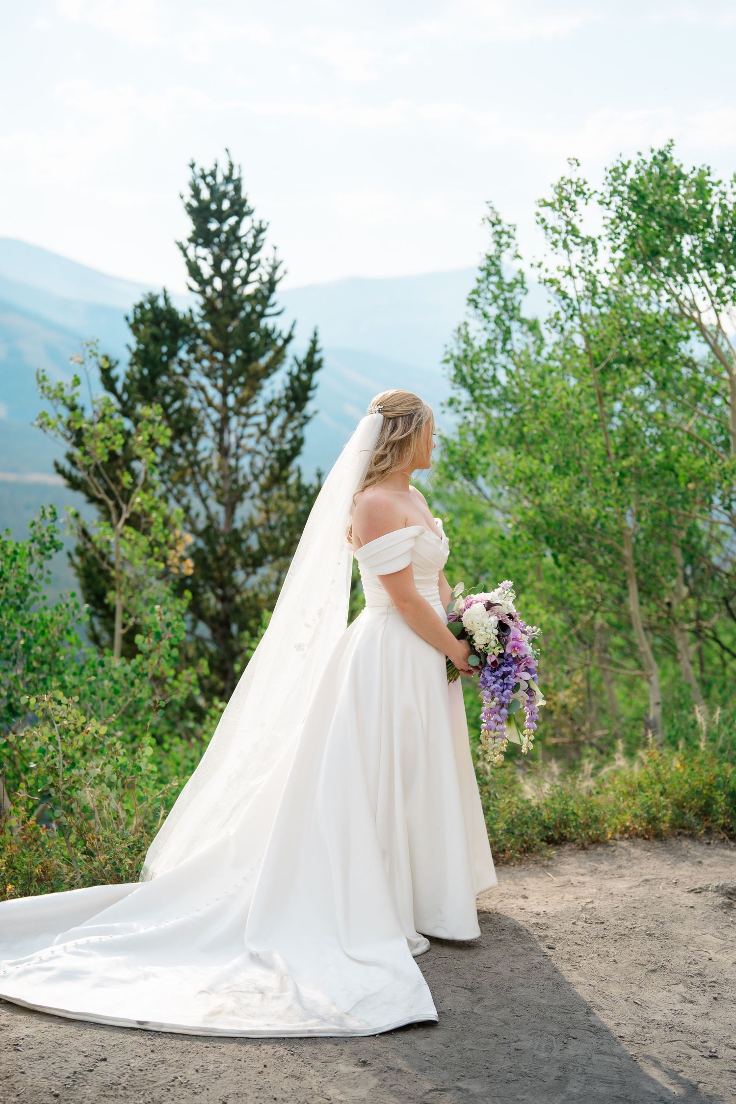 A bride in an elegant off-shoulder white gown holds a purple bouquet while standing on a mountain overlook during breckenridge micro wedding