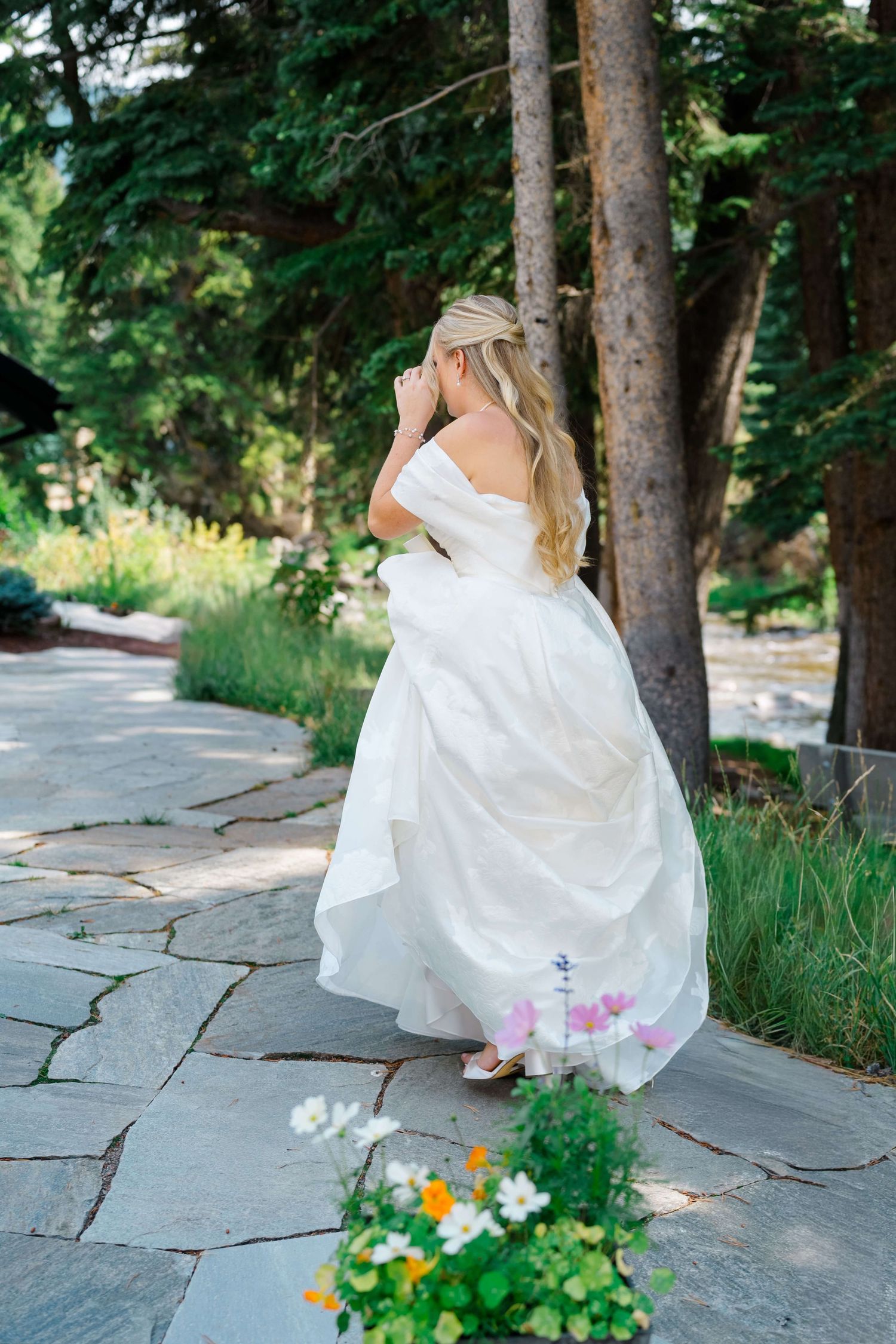 A bride in a flowing white wedding dress walks along a stone path surrounded by evergreen trees and colorful flowers at vail wedding.