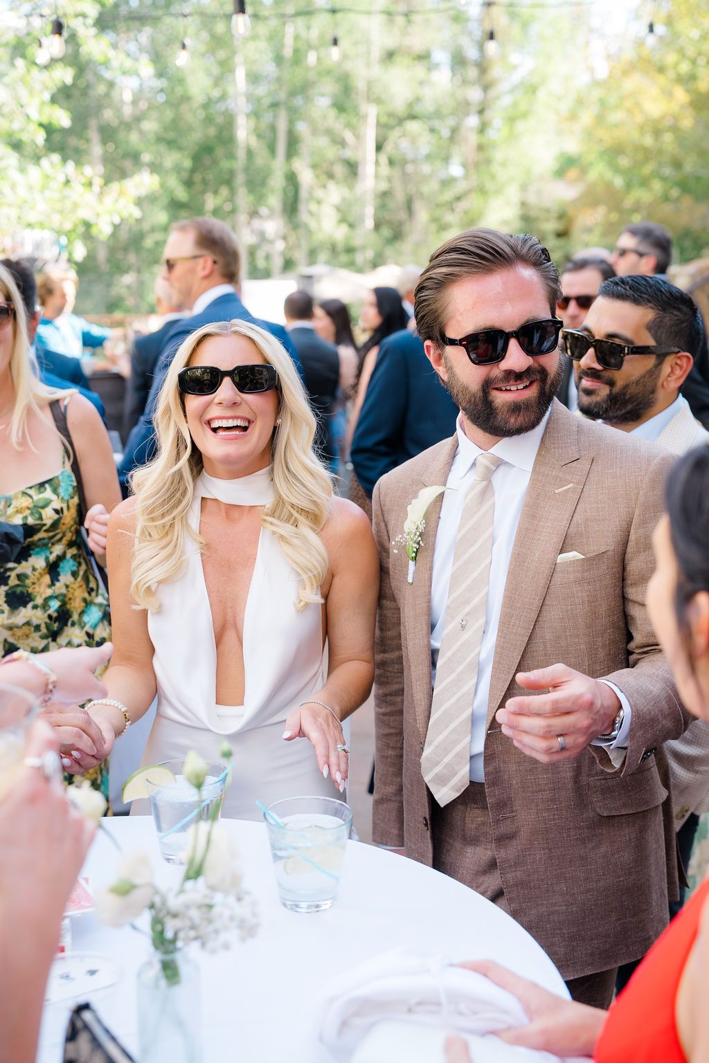 Smiling couple in sunglasses at outdoor wedding reception, woman in white dress, man in tan suit with boutonniere.
