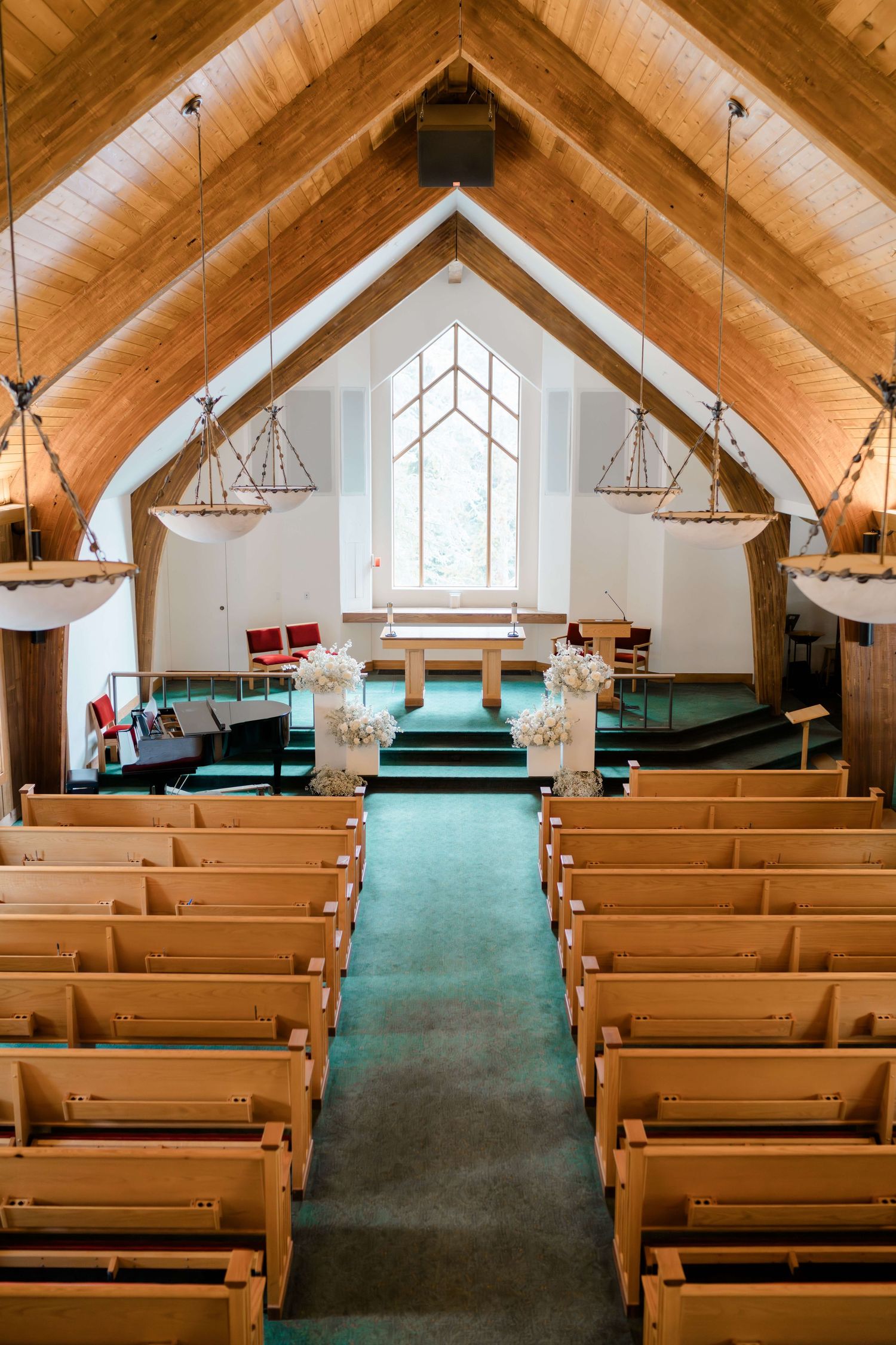 Wooden A-frame chapel interior with teal carpet aisle, pine pews, and floral arrangements leading to a bright altar.