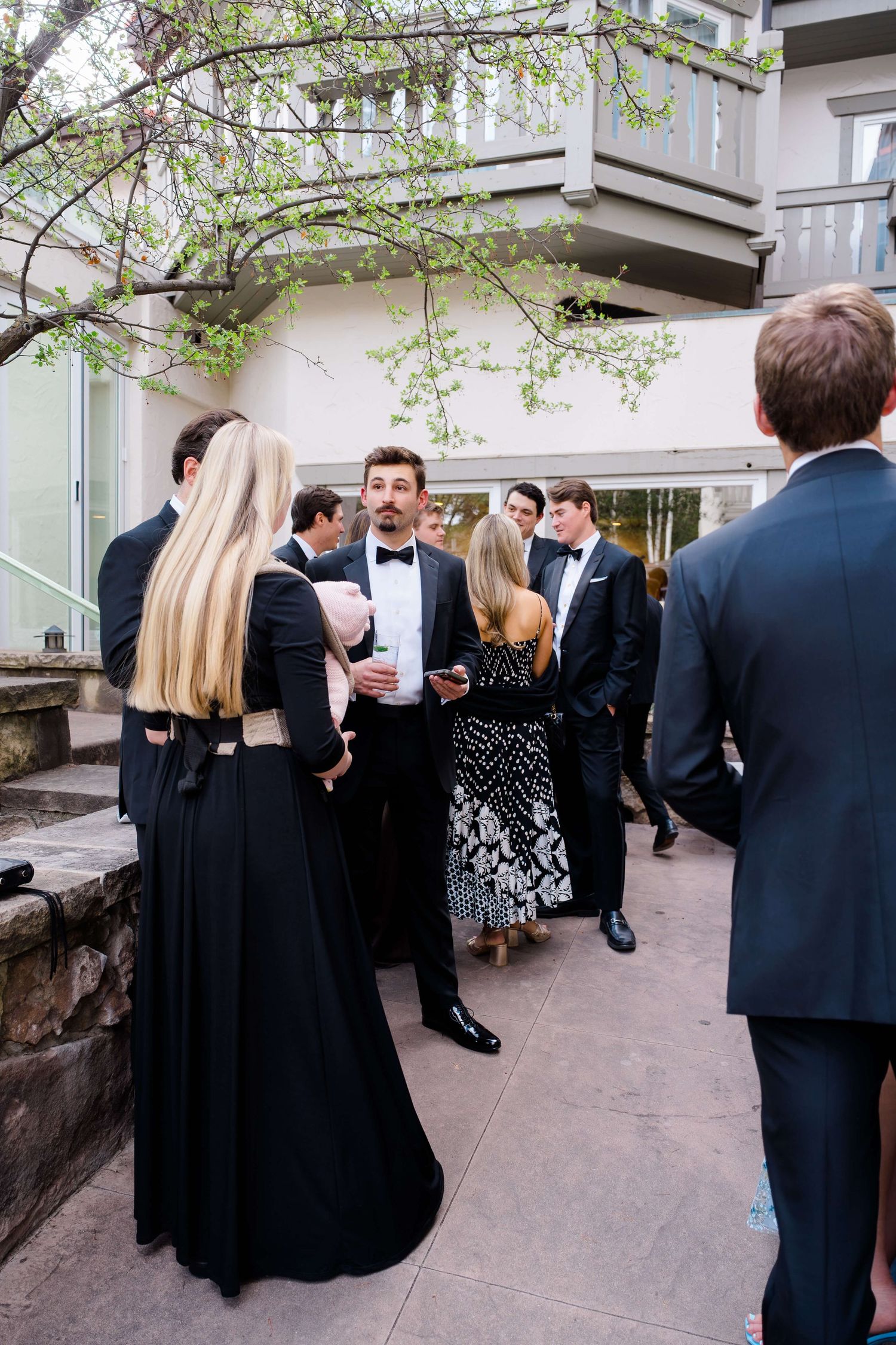 Elegantly dressed guests in black tie attire mingle outdoors at a formal wedding cocktail hour.