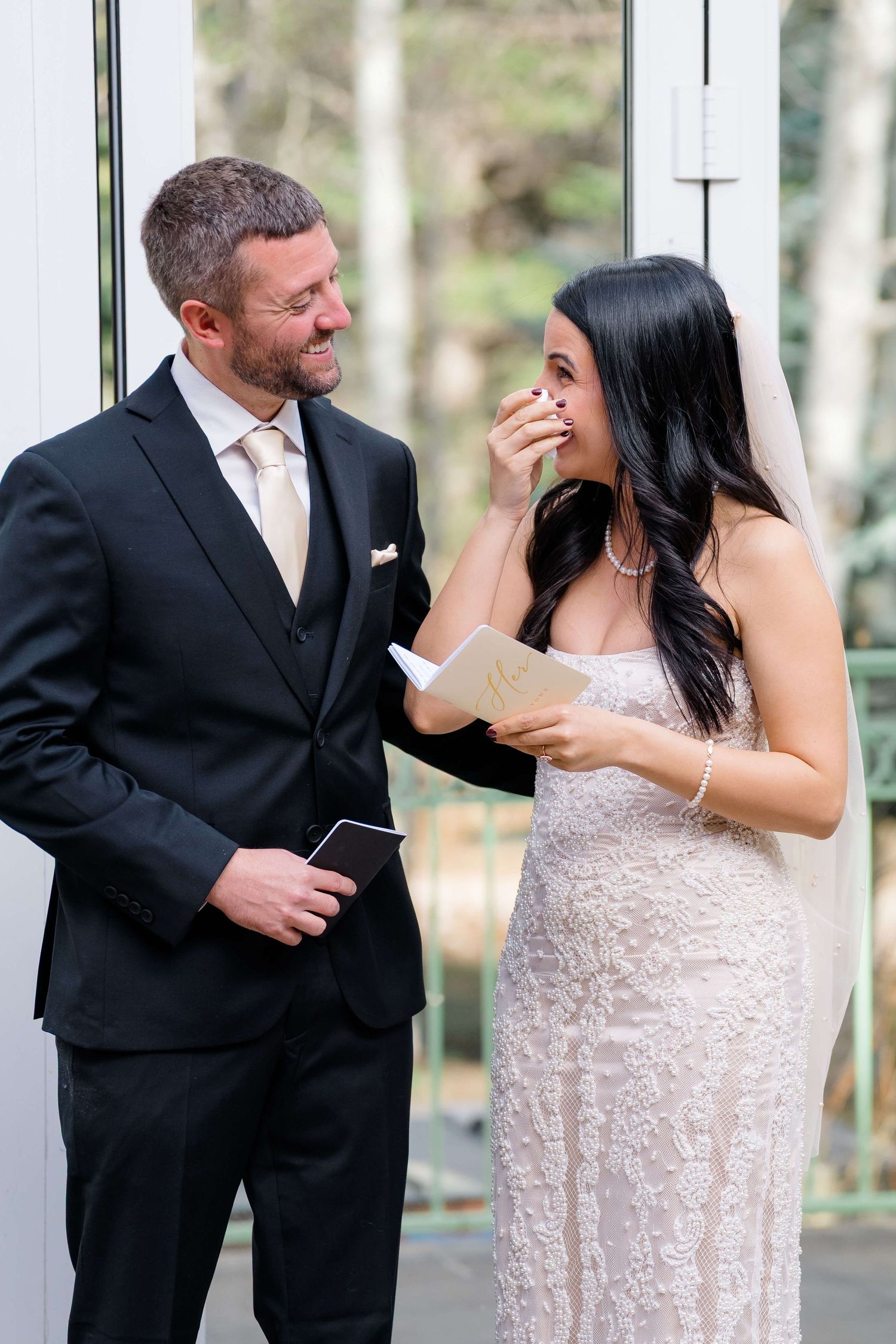 Bride wipes tears while groom smiles, exchanging vows during an emotional wedding ceremony.