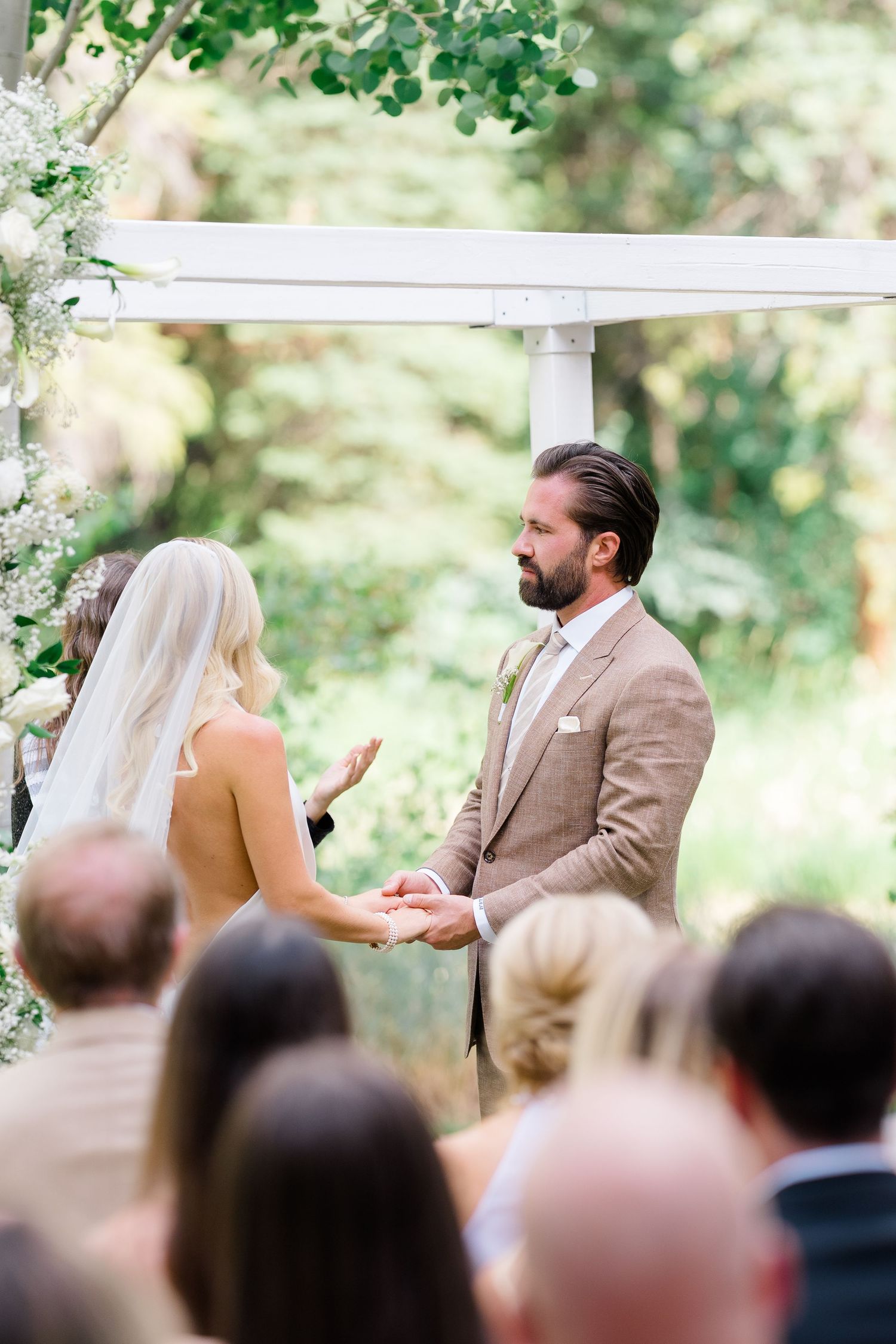 Bride and groom exchange vows outdoors under a white floral arbor, surrounded by lush greenery and wedding guests.