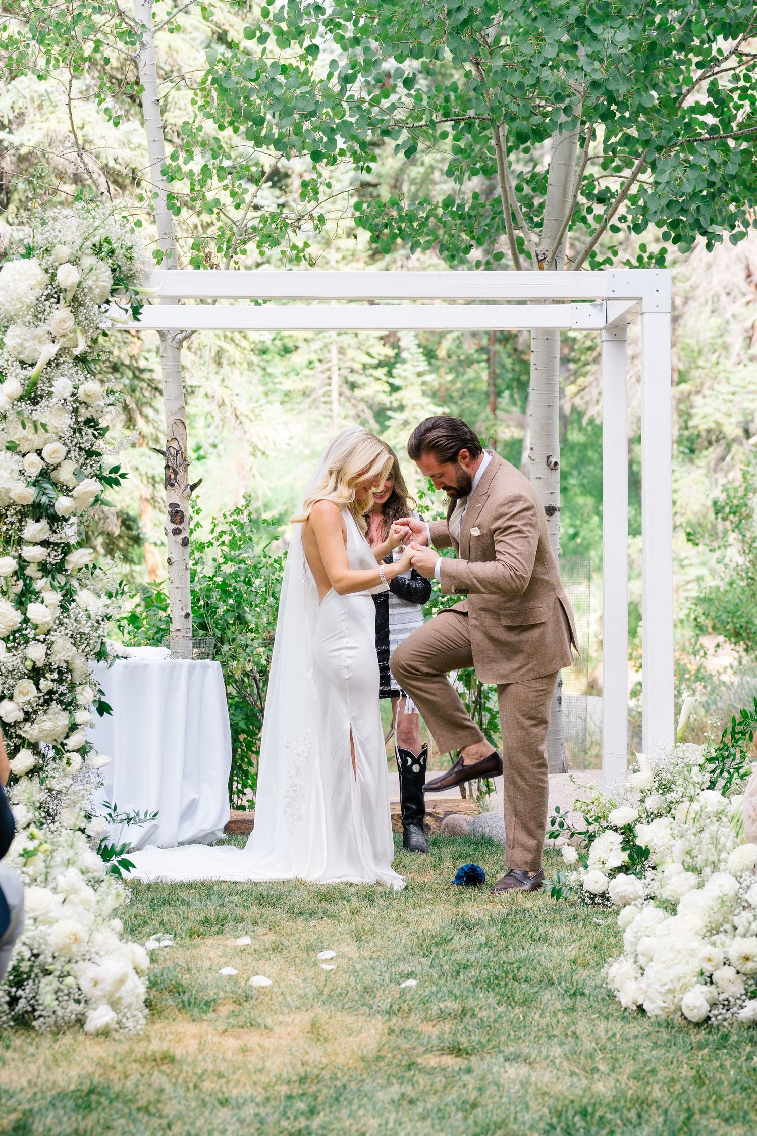 Outdoor wedding ceremony with bride and groom exchanging rings under a white floral arch surrounded by lush greenery.