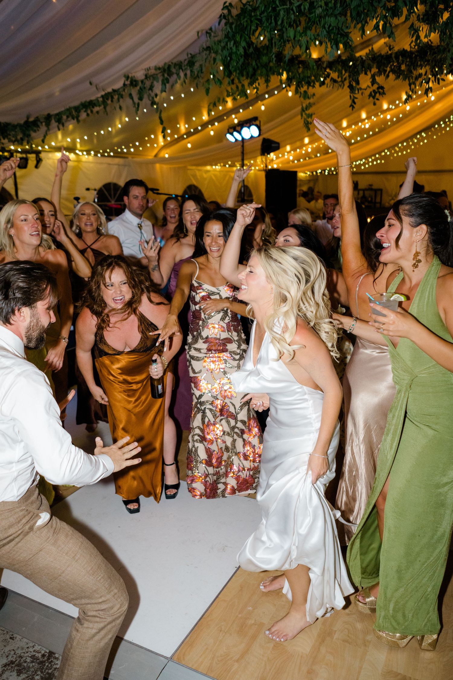 Bride in white satin dress dances joyfully with guests at a wedding reception under warm string lights.