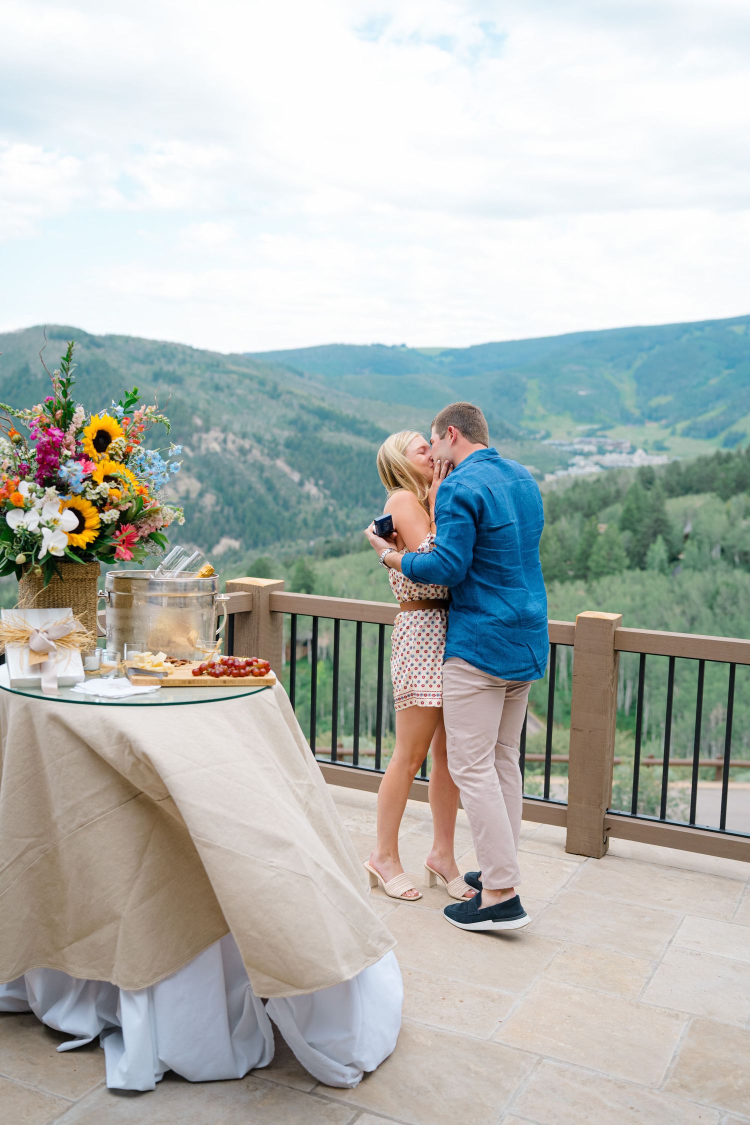 guy standing with a ring box as he kisses his fiance and is so excited as they are now engaged - background is mountains and a floral set up