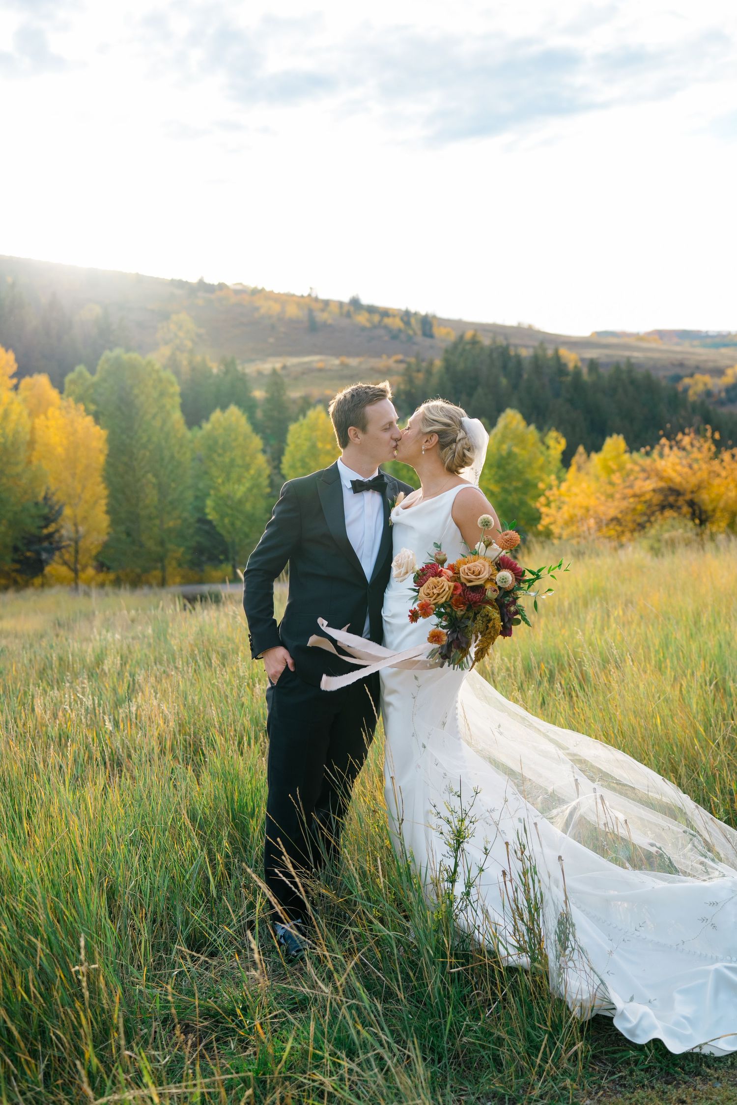 A romantic wedding couple embraces in a golden autumn meadow with mountains and colorful fall foliage in the background.