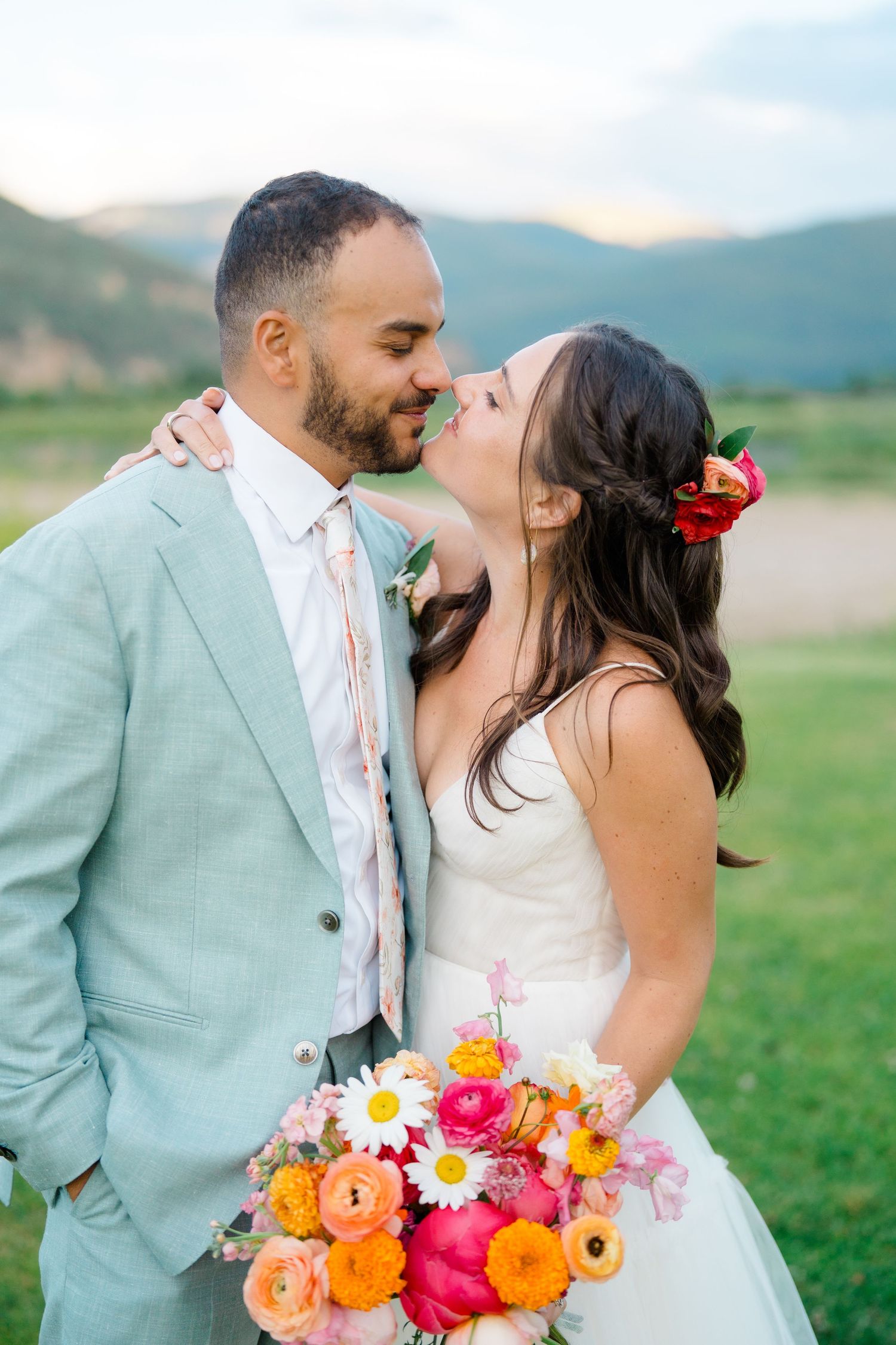 A couple shares romantic kisses in a scenic outdoor setting with vibrant floral bouquets in shades of pink and orange.