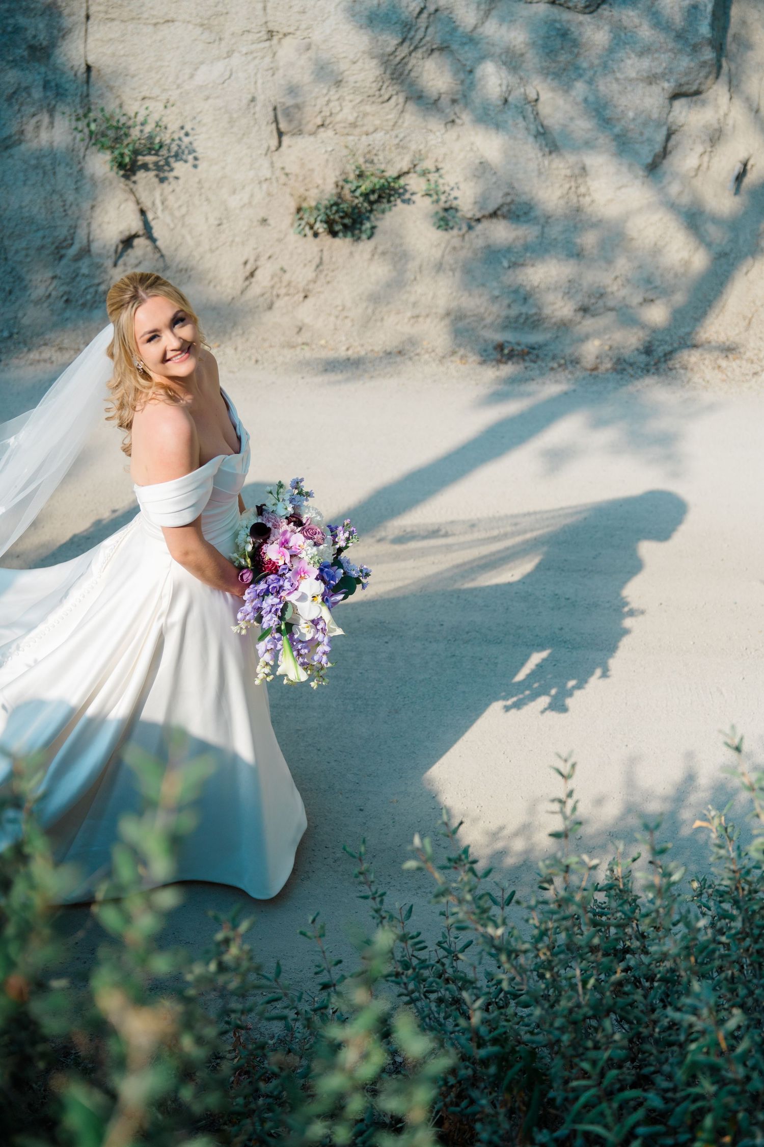 A bride in an elegant white dress twirls in dappled sunlight against a stone wall with purple flowers.