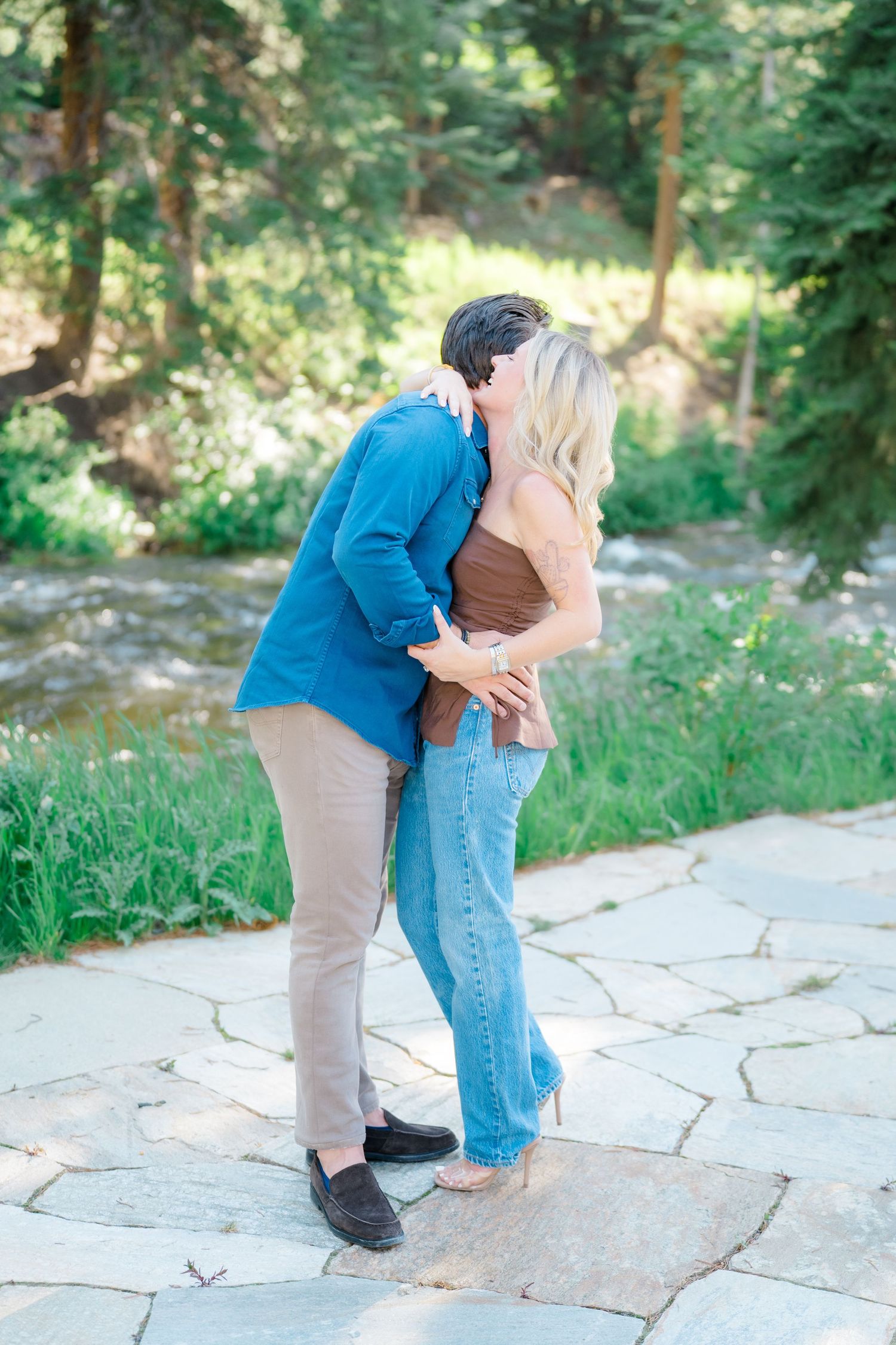 A couple shares a romantic moment on stone steps surrounded by lush greenery and trees in a natural outdoor setting.