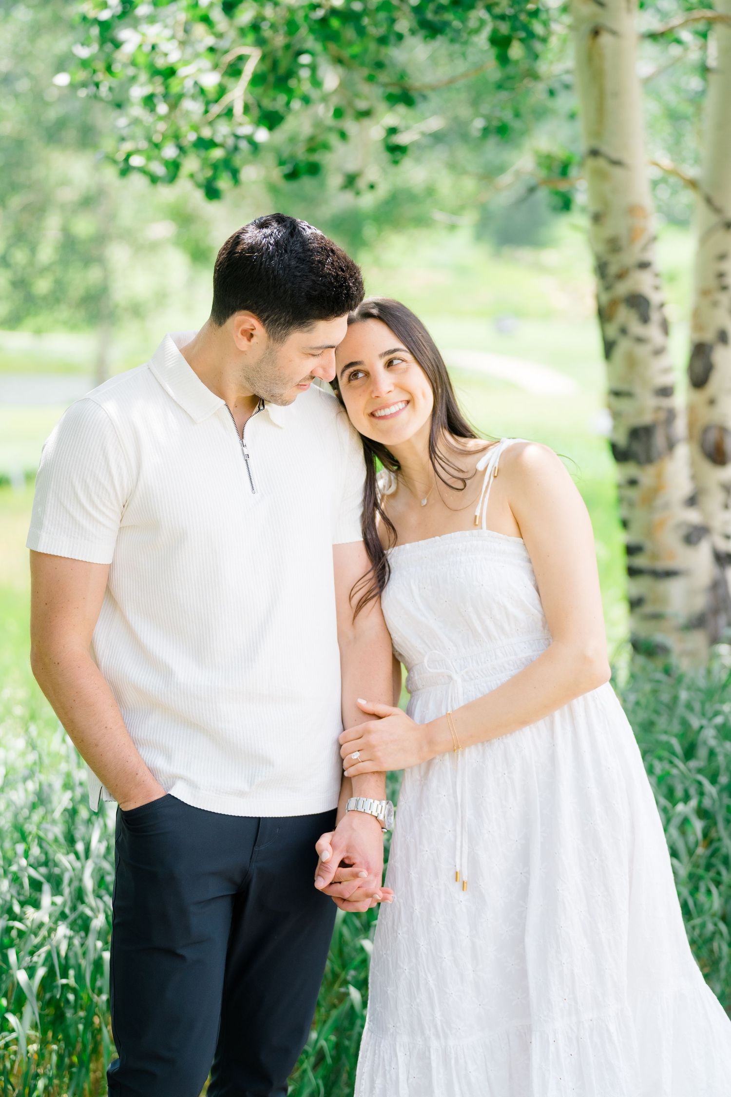 A romantic couple in white clothing shares an intimate moment among birch trees and wildflowers.