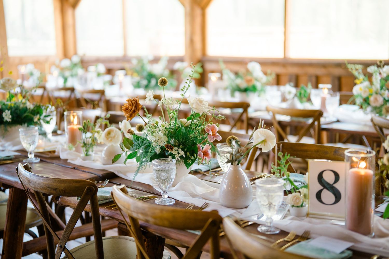 Elegant rustic wedding reception tables set with white linens, greenery centerpieces and wooden cross-back chairs.