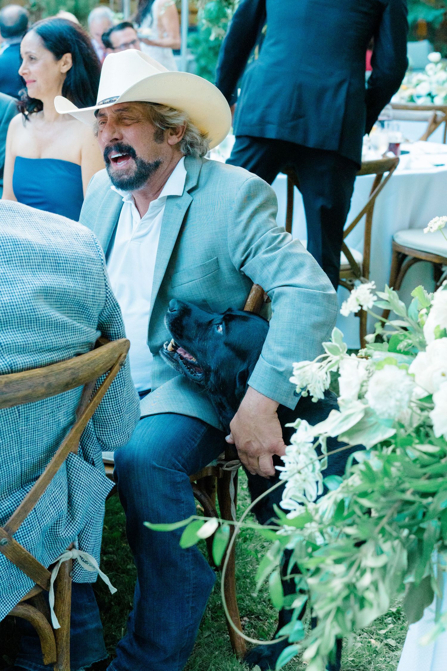 Wedding guests in cowboy hats sit in wooden chairs surrounded by white floral arrangements at an outdoor ceremony.