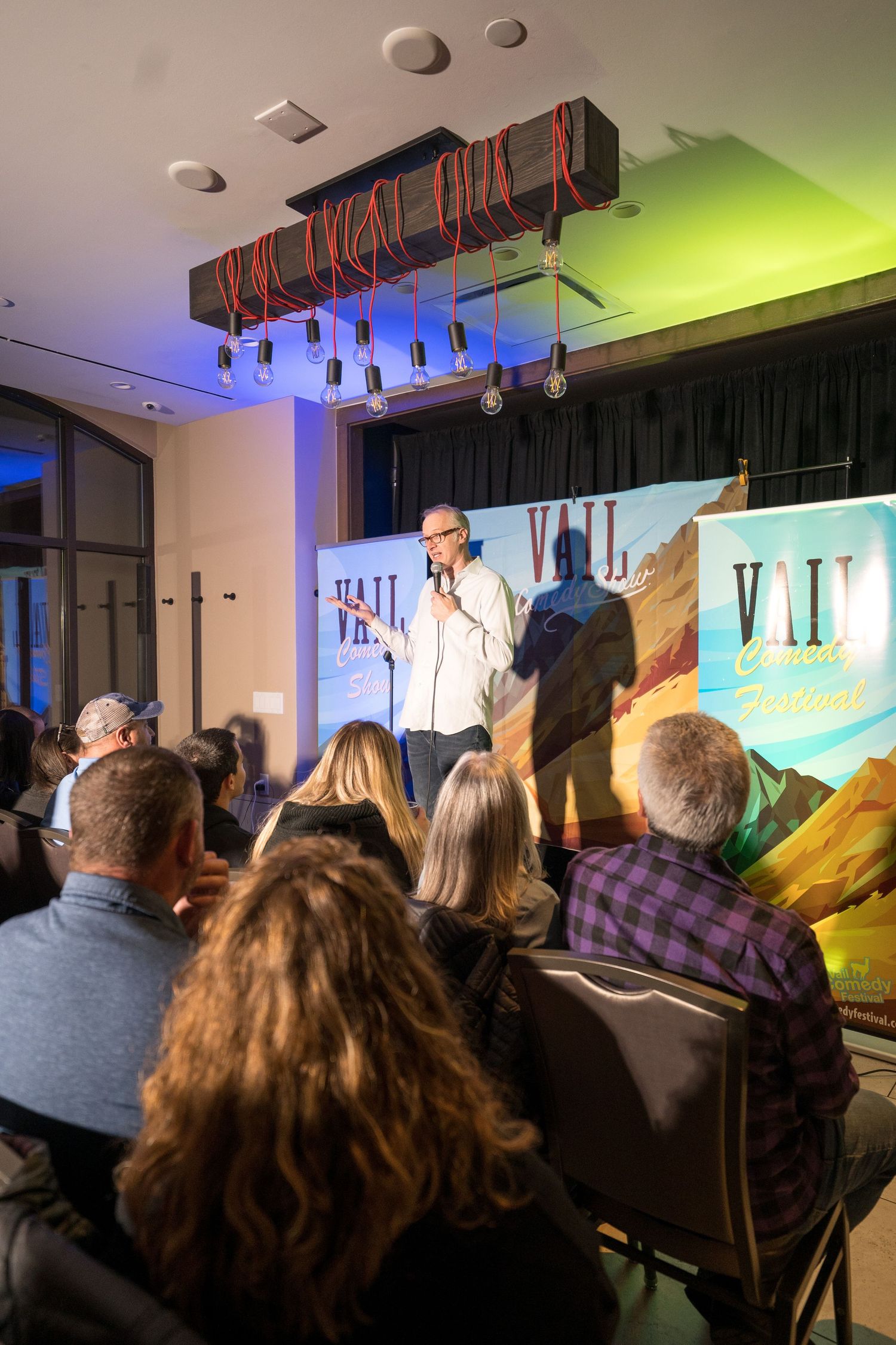 A performer on stage at an event with a mountain backdrop banner and distinctive ceiling lighting.