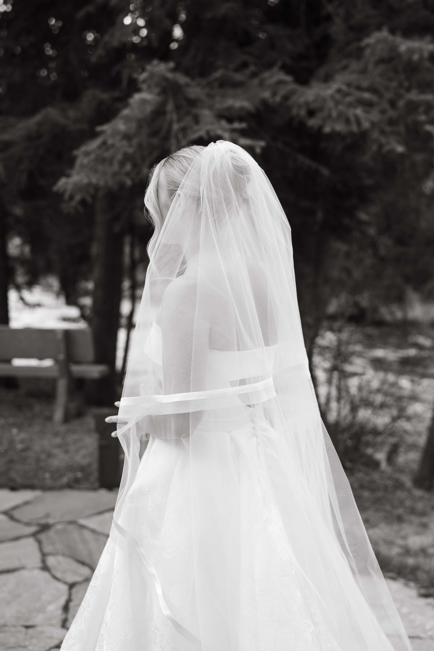 Black and white photo of a bride in a flowing white gown and veil, standing outdoors among tree at Vail Sonnenalp Wedding.