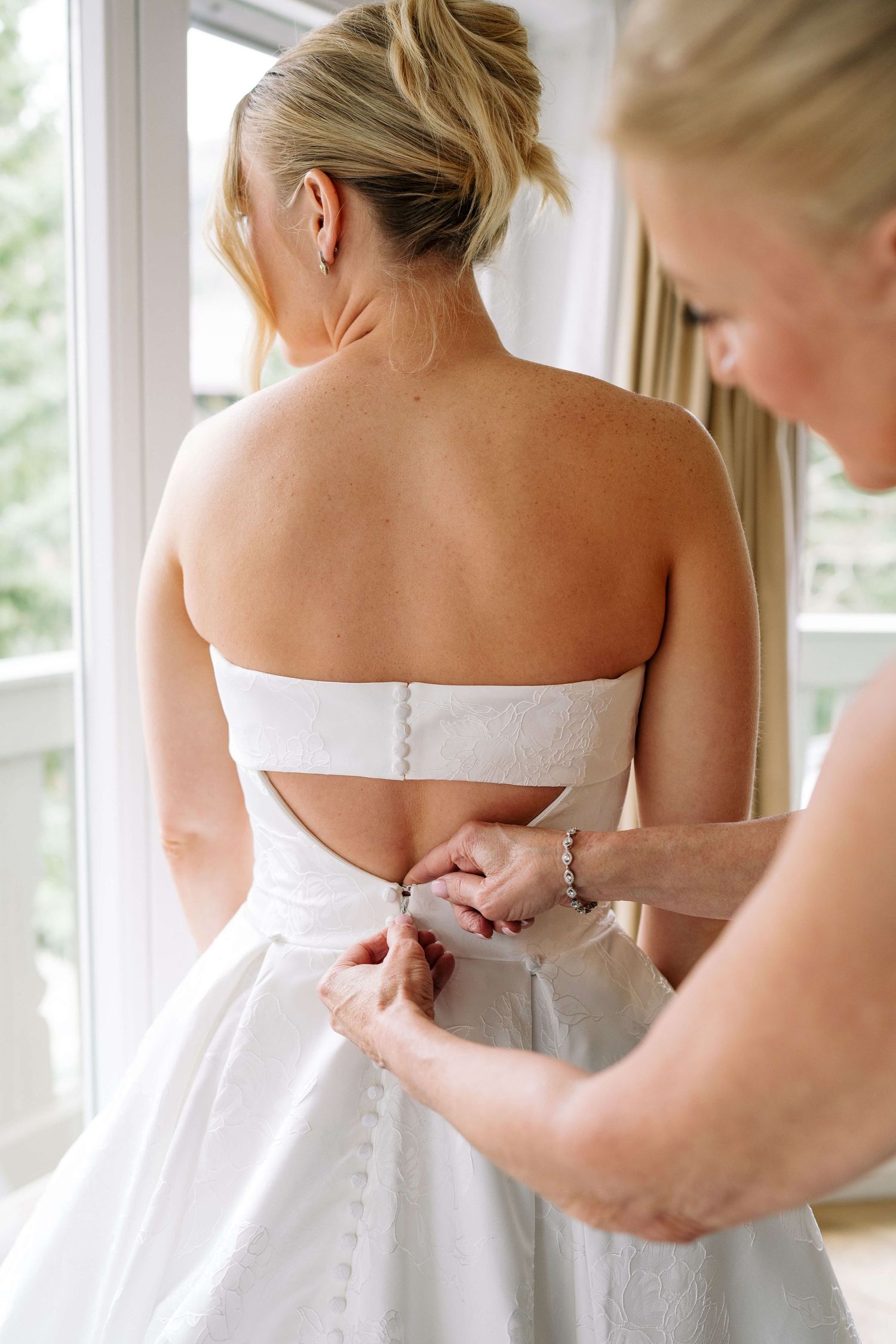 Bride getting her strapless white wedding dress buttoned up from behind by a helper near a bright window at Vail Sonnenalp Wedding.
